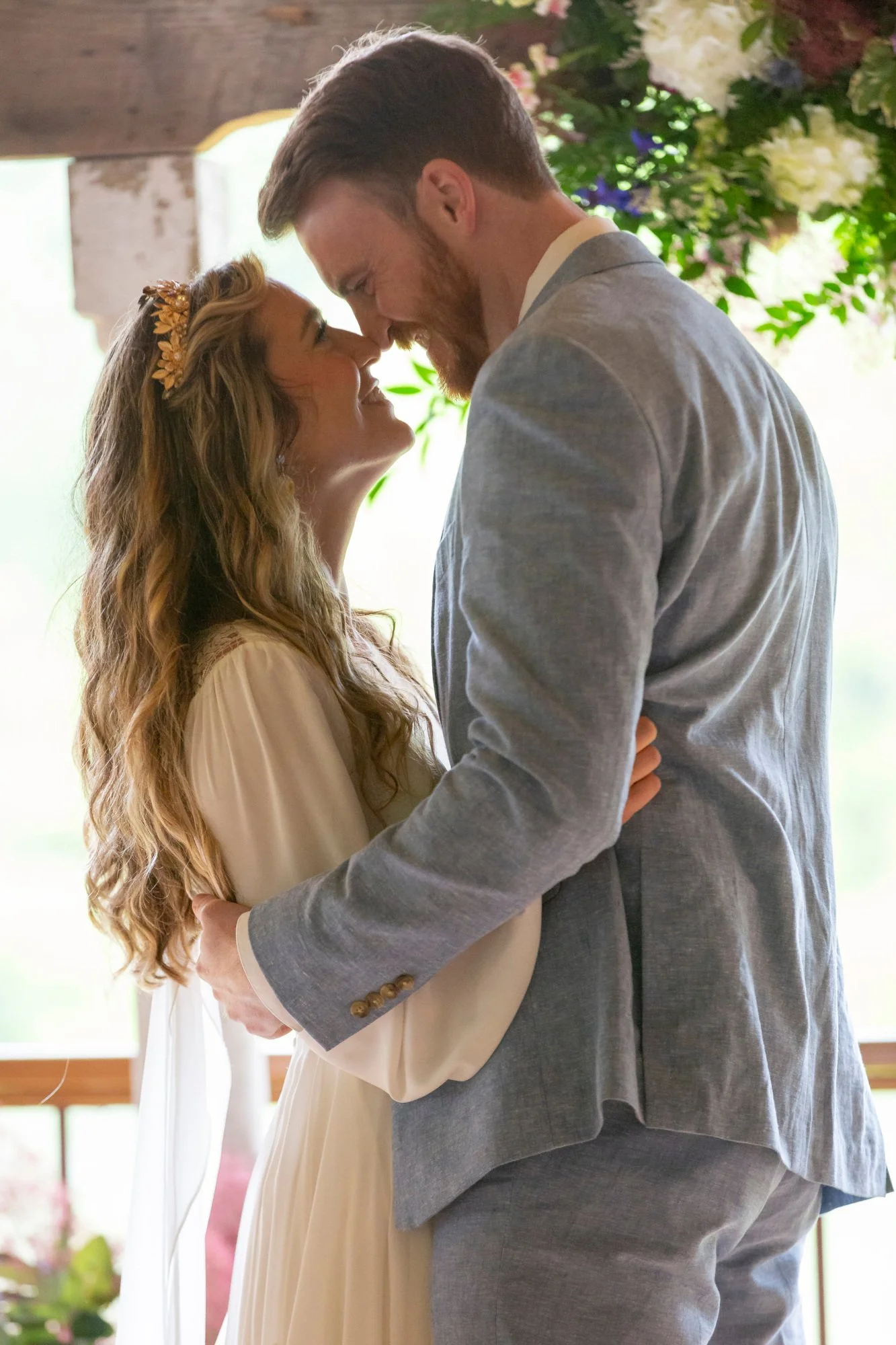 A man and woman are standing close together, touching foreheads and smiling during a wedding or romantic ceremony in a well-lit indoor space with flowers in the background.
