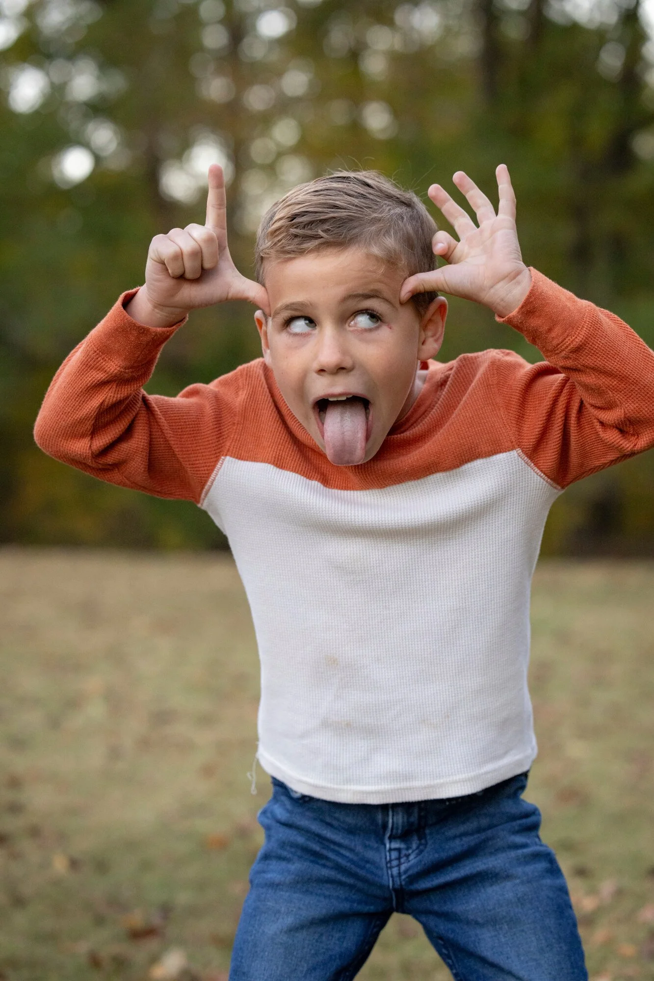 A young boy standing outdoors, wearing a long-sleeve orange and white shirt and blue jeans, sticking out his tongue and making silly hand gestures near his head, with a blurred background of trees.