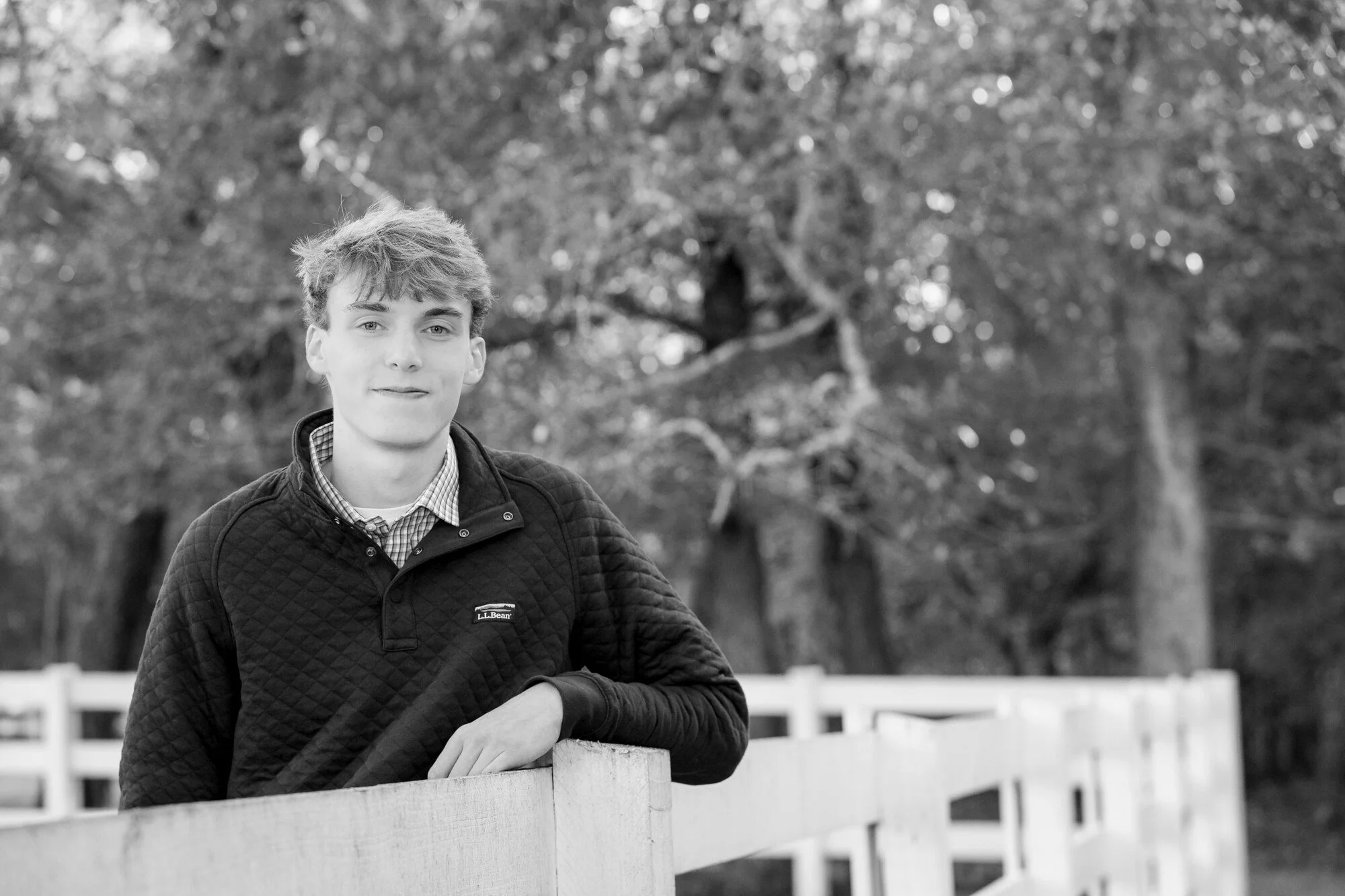 A young man with light hair and fair skin leans on a white picket fence outdoors, wearing a dark quilted jacket and a checkered shirt, with a background of trees.
