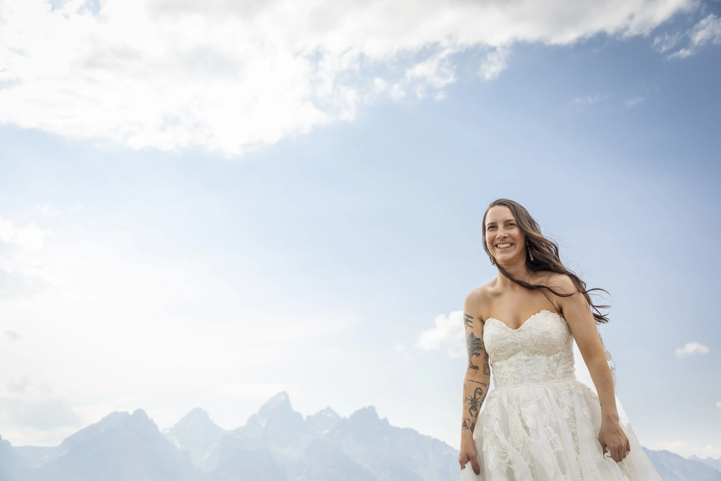 A woman with long brown hair wearing a white strapless wedding dress, smiling outdoors with mountains and a cloudy sky in the background.