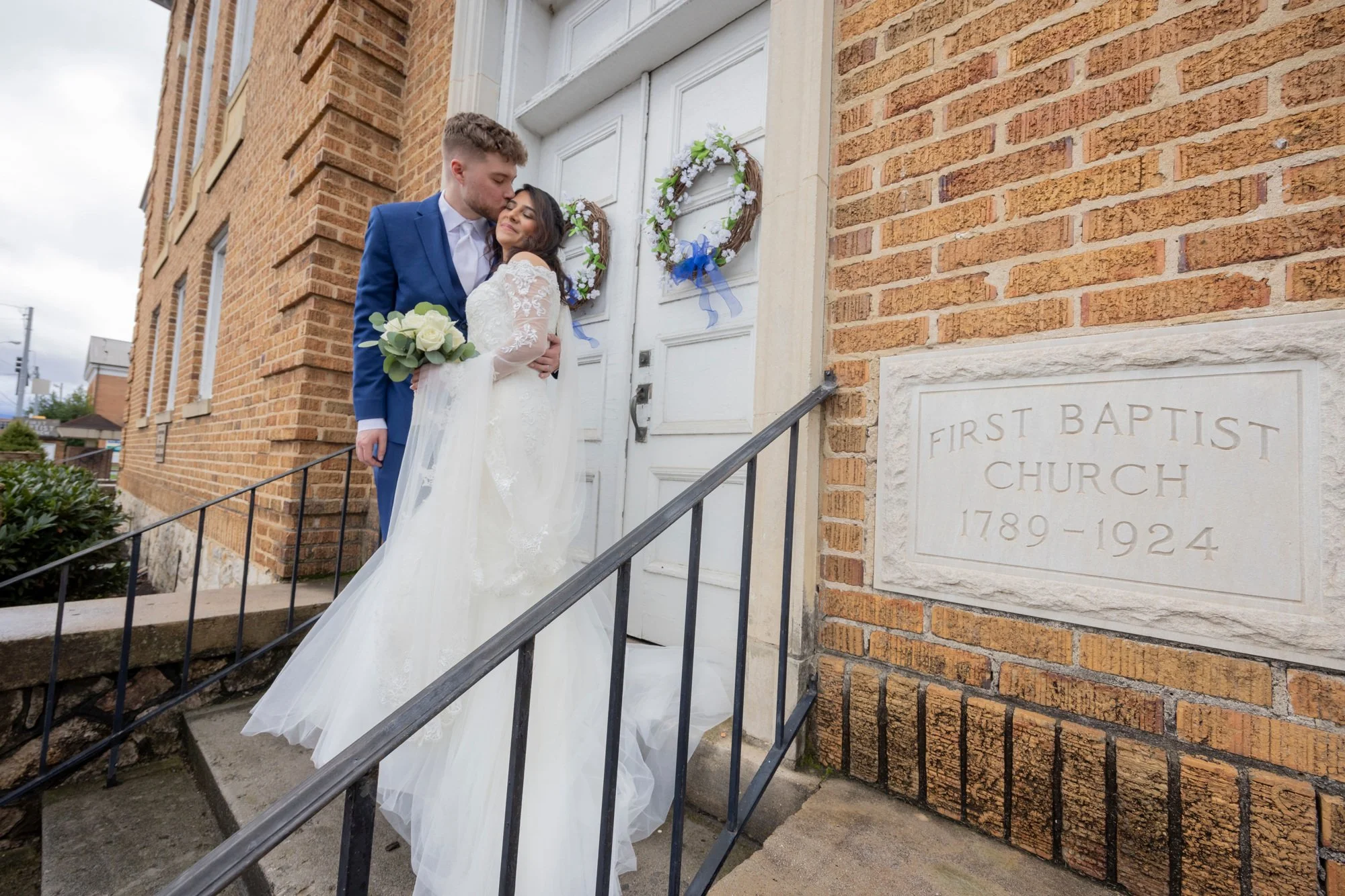 A bride and groom standing at the door of a church, with the groom kissing the bride on her forehead. The bride is holding a bouquet of white roses and wearing a white lace wedding dress. The church door has wedding wreaths with blue ribbons, and a s