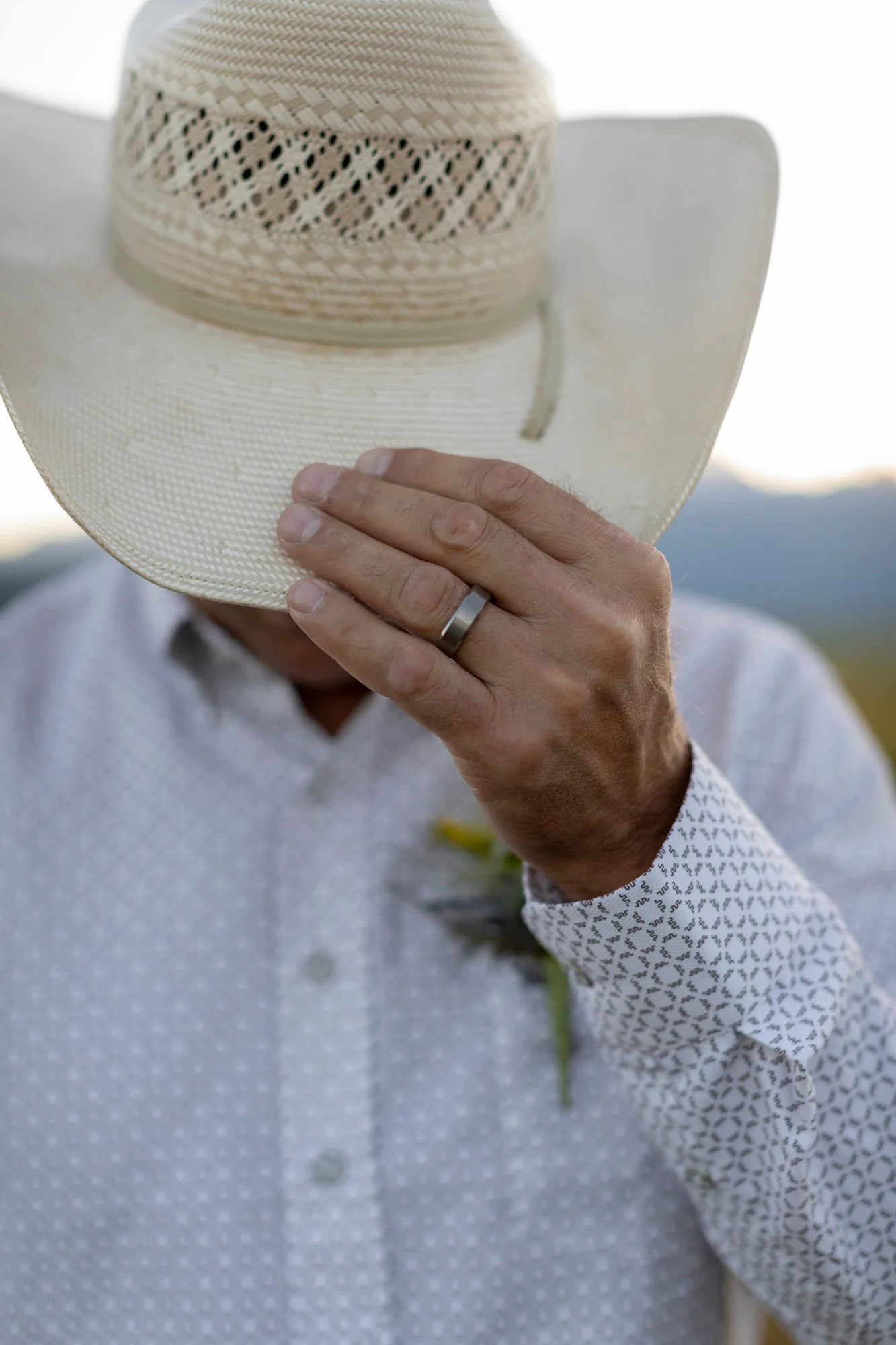 A person holding a large, off-white sun hat with a decorative band, wearing a light-colored, patterned button-up shirt with rolled-up sleeves.