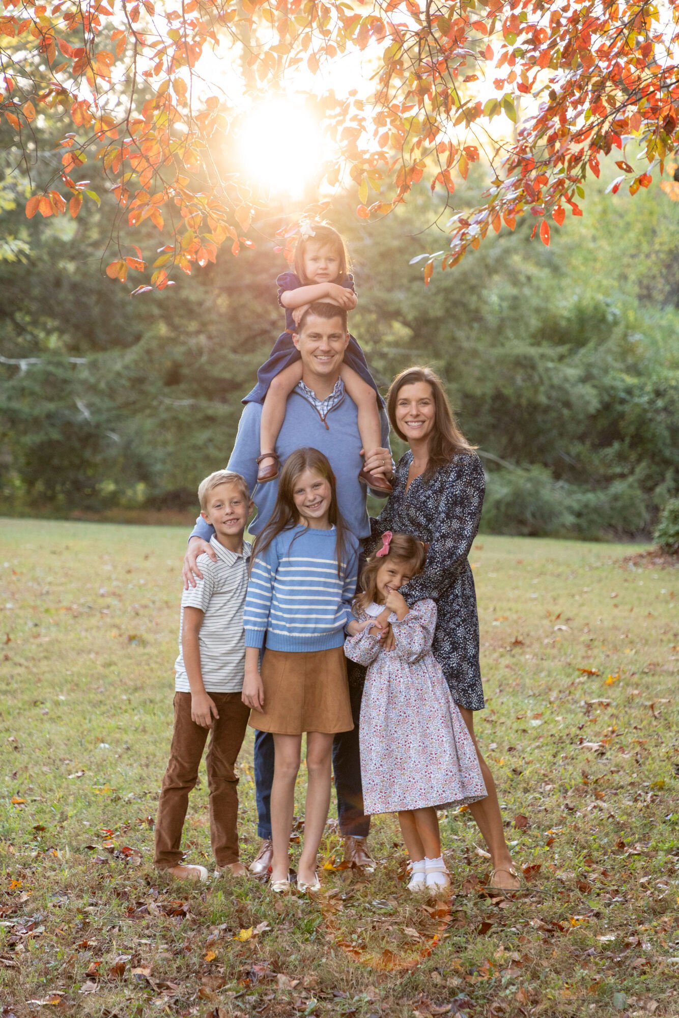 A family of seven outdoors in a park with autumn leaves. A man carries a young girl on his shoulders, and a woman stands beside him. Three children stand in front, and another girl is in the woman's arms. The sunlight filters through tree branches wi