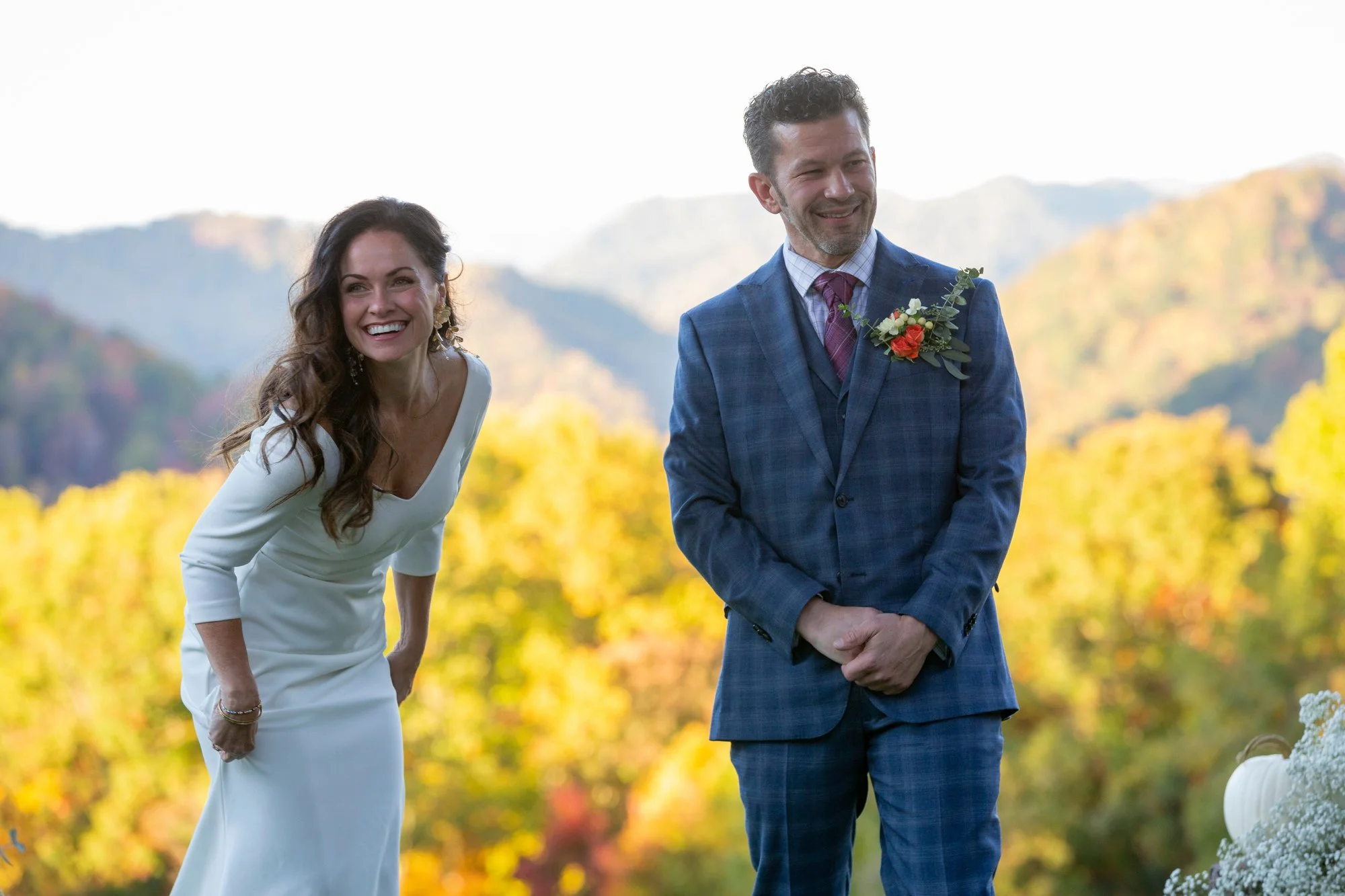A smiling woman in a white dress leaning forward and a smiling man in a suit with a boutonniere standing outdoors amidst colorful fall foliage and distant mountains.