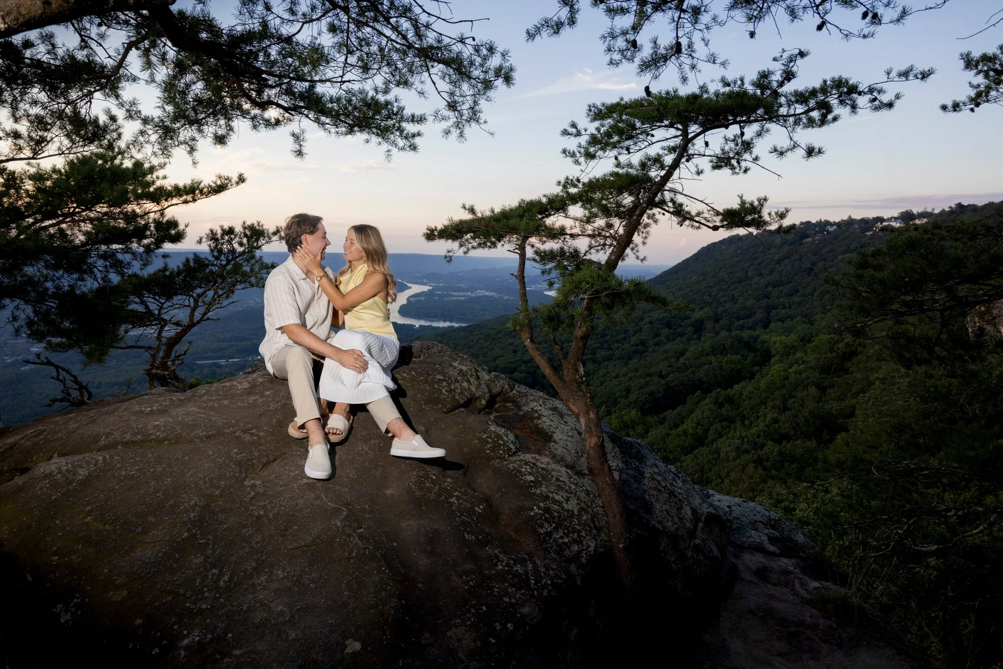 A couple sitting on a large rock on a mountain with a scenic view of a river and green valley below, surrounded by trees, during sunset.