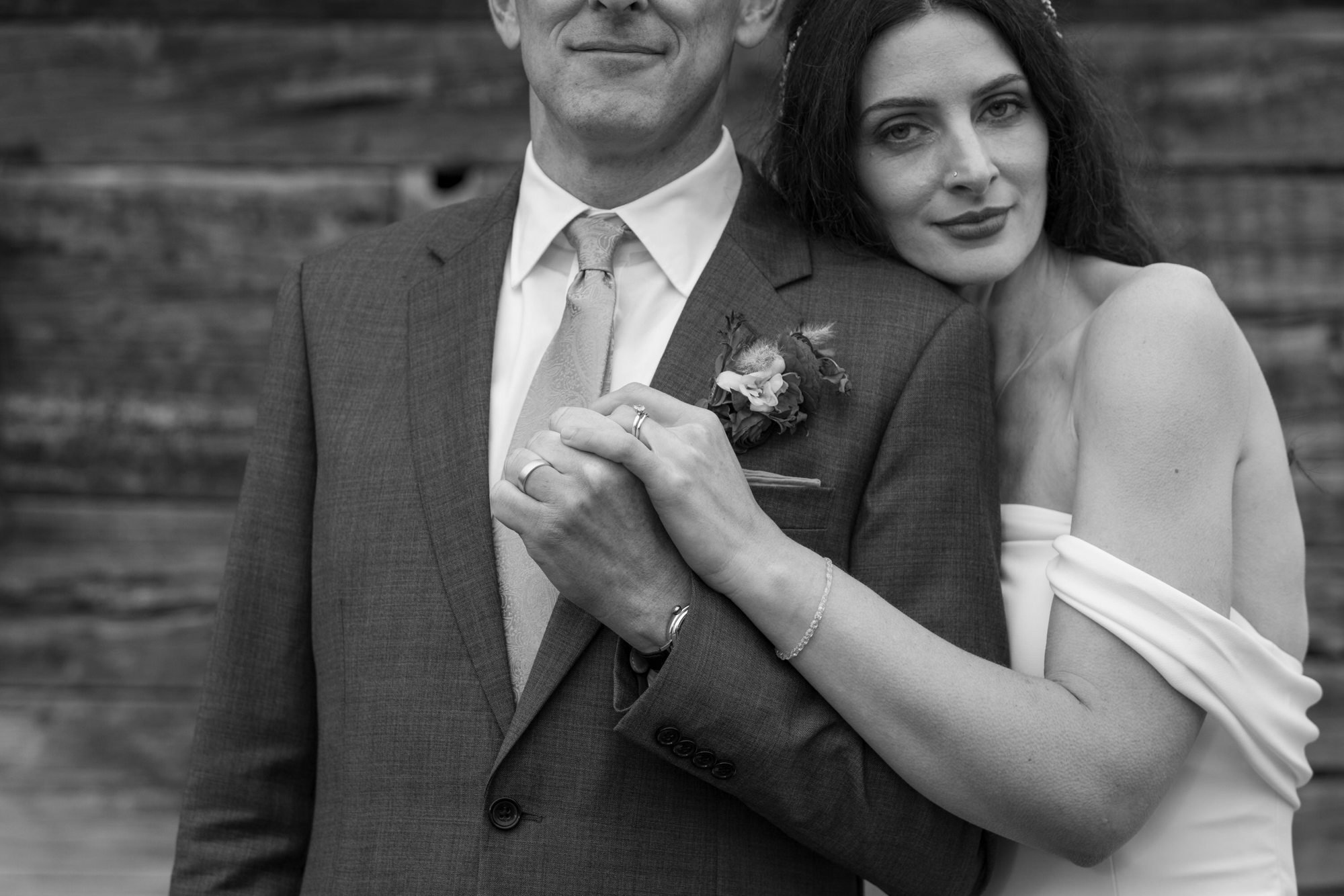 Black and white photo of a couple, with the woman resting her head on the man's shoulder, both smiling gently. The man is dressed in a suit with a tie and boutonniere, and the woman is wearing a dress and jewelry. They are celebrating a wedding or sp