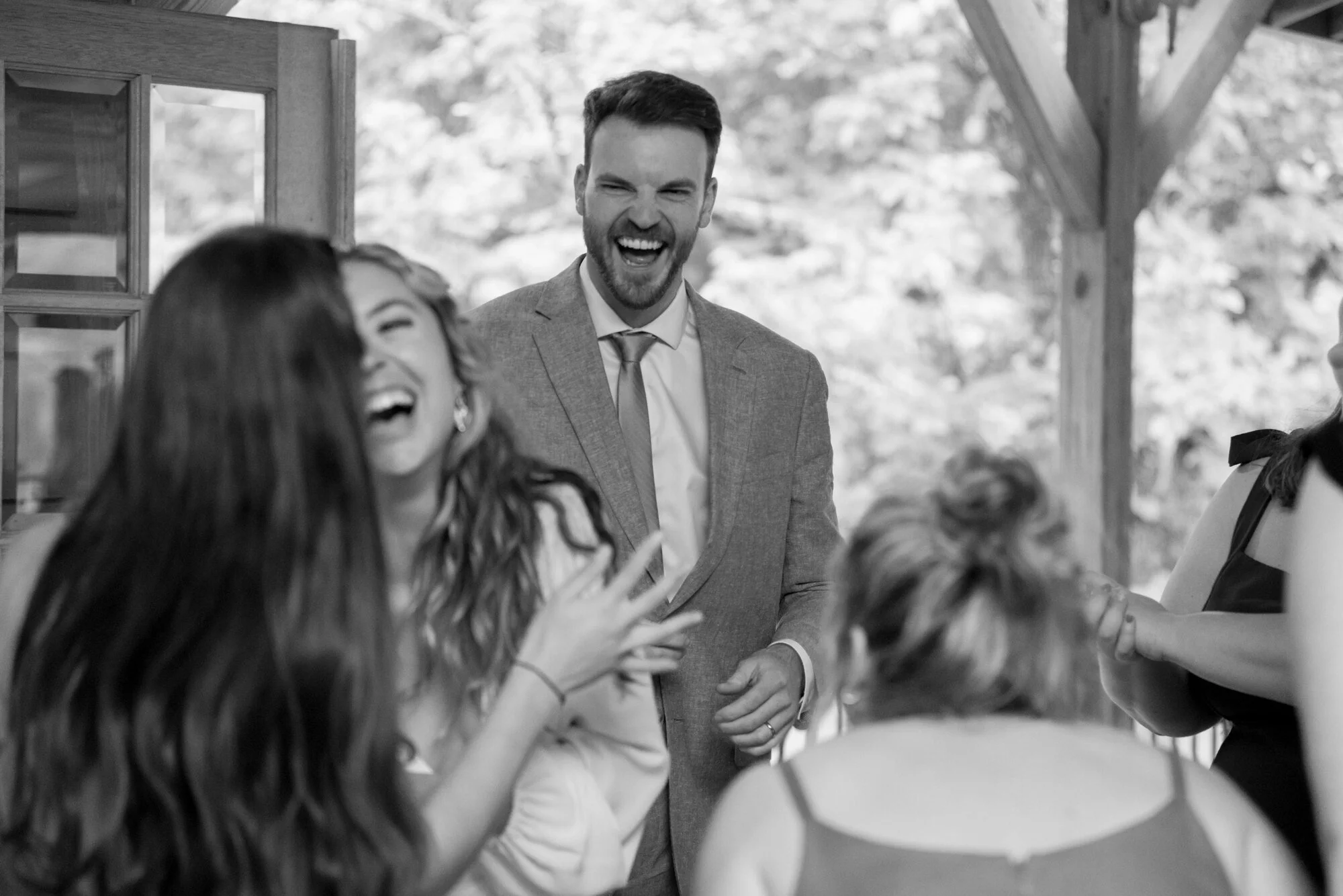 A group of people at an outdoor wedding reception, with a man in a suit smiling and laughing, surrounded by women who are also smiling and chatting.