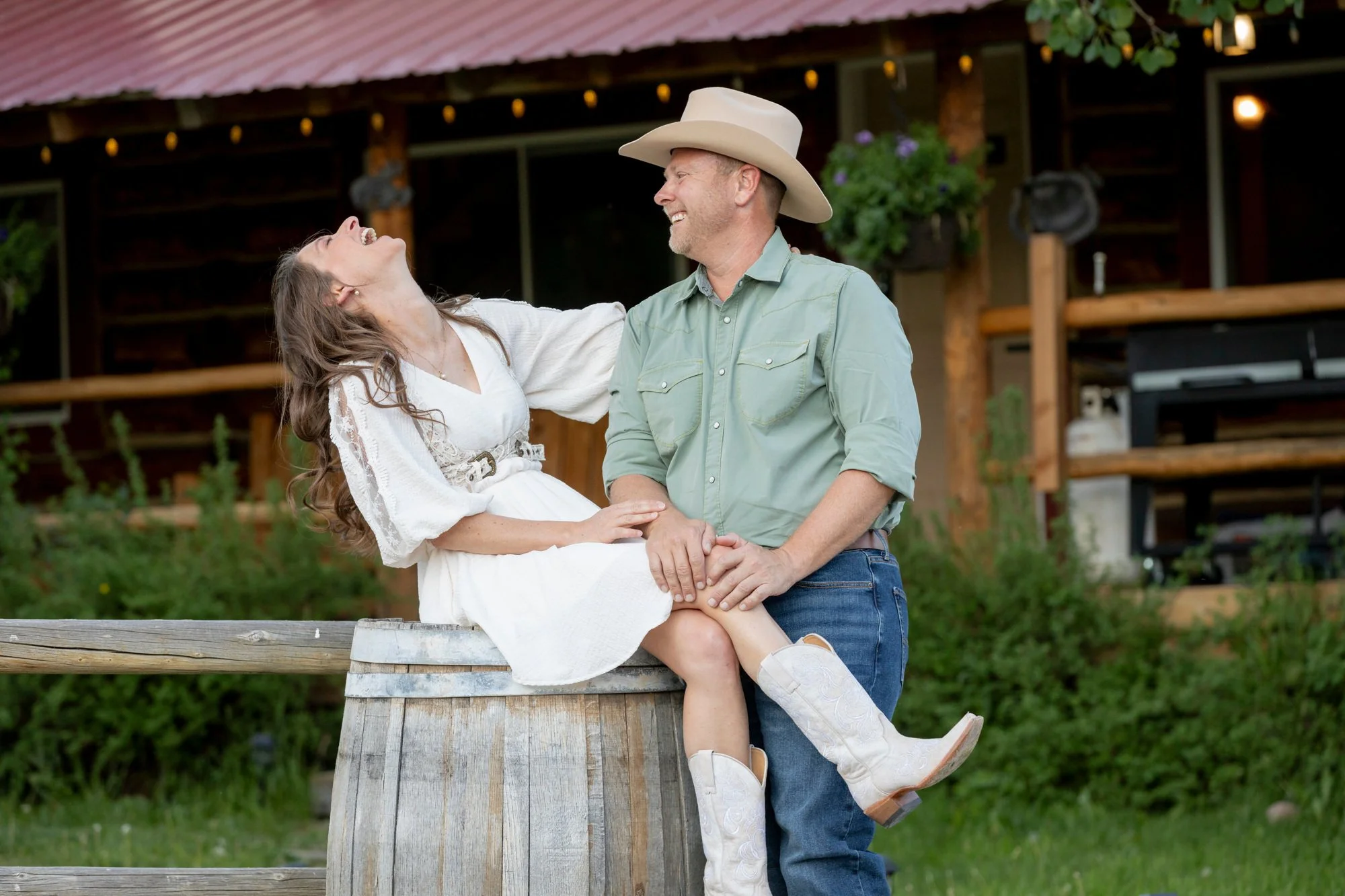 A couple happy and laughing, the woman sitting on a wooden barrel, the man standing and holding her, outside in front of a rustic building with a red roof and hanging flower basket.