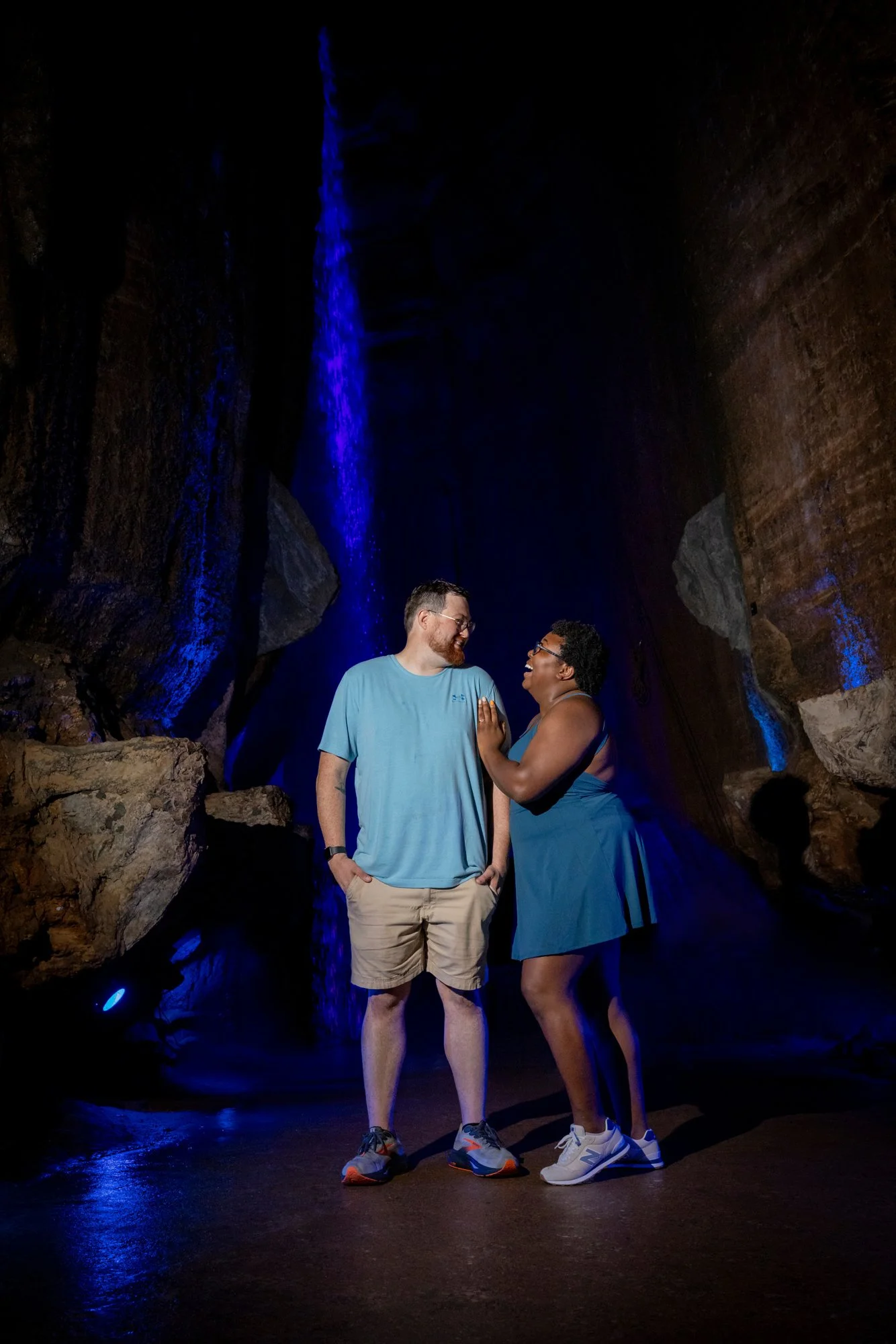 A man and a woman standing close together inside a dark cavern with rocks and a waterfall illuminated in blue light. They are smiling, facing each other affectionately.