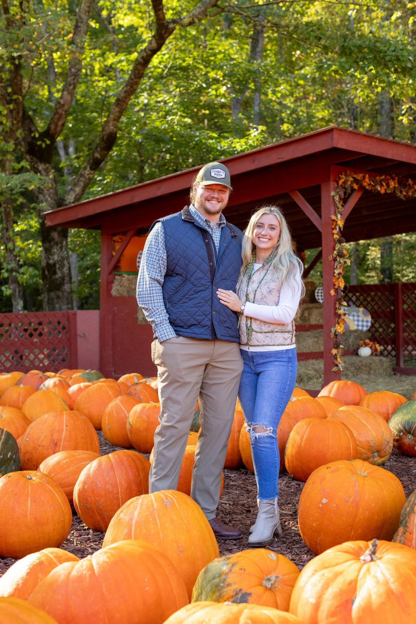 A smiling man and woman standing together among pumpkins in an outdoor pumpkin patch during fall, with trees and a red wooden shelter decorated with autumn leaves in the background.