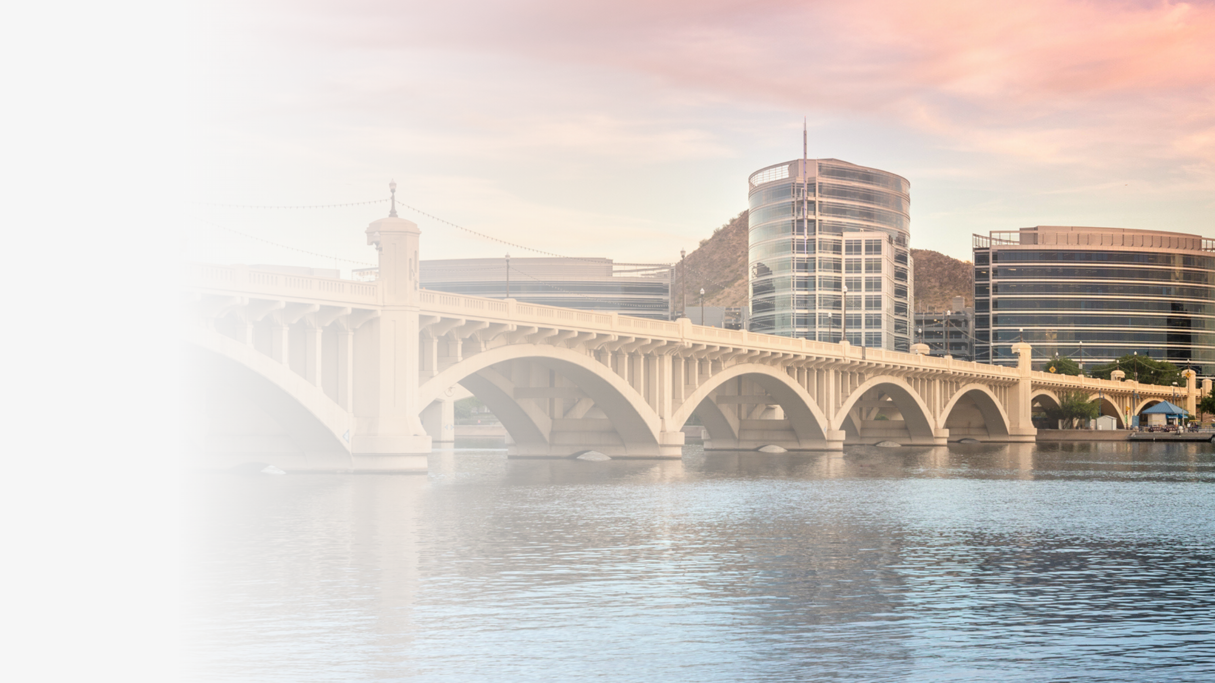 Cityscape with modern glass buildings, a bridge over water, and hills in the background during sunset