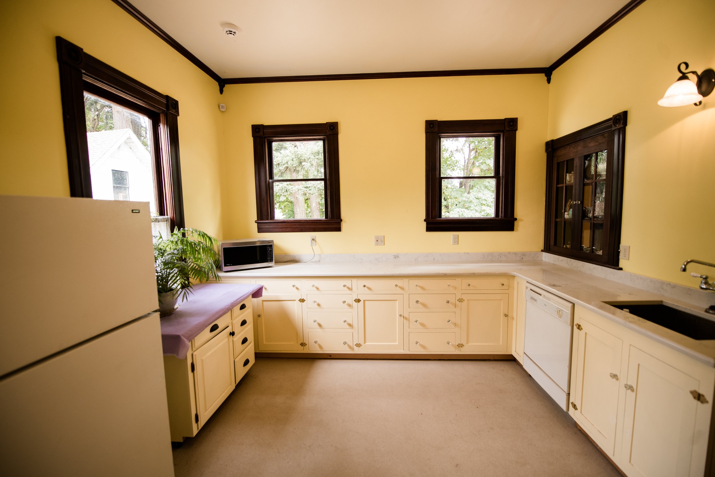 The kitchen at the Brown House Event Center  with yellow walls, dark wood trim around windows and cabinets, three windows showing trees outside, a microwave on the counter, a plant, and a white refrigerator.