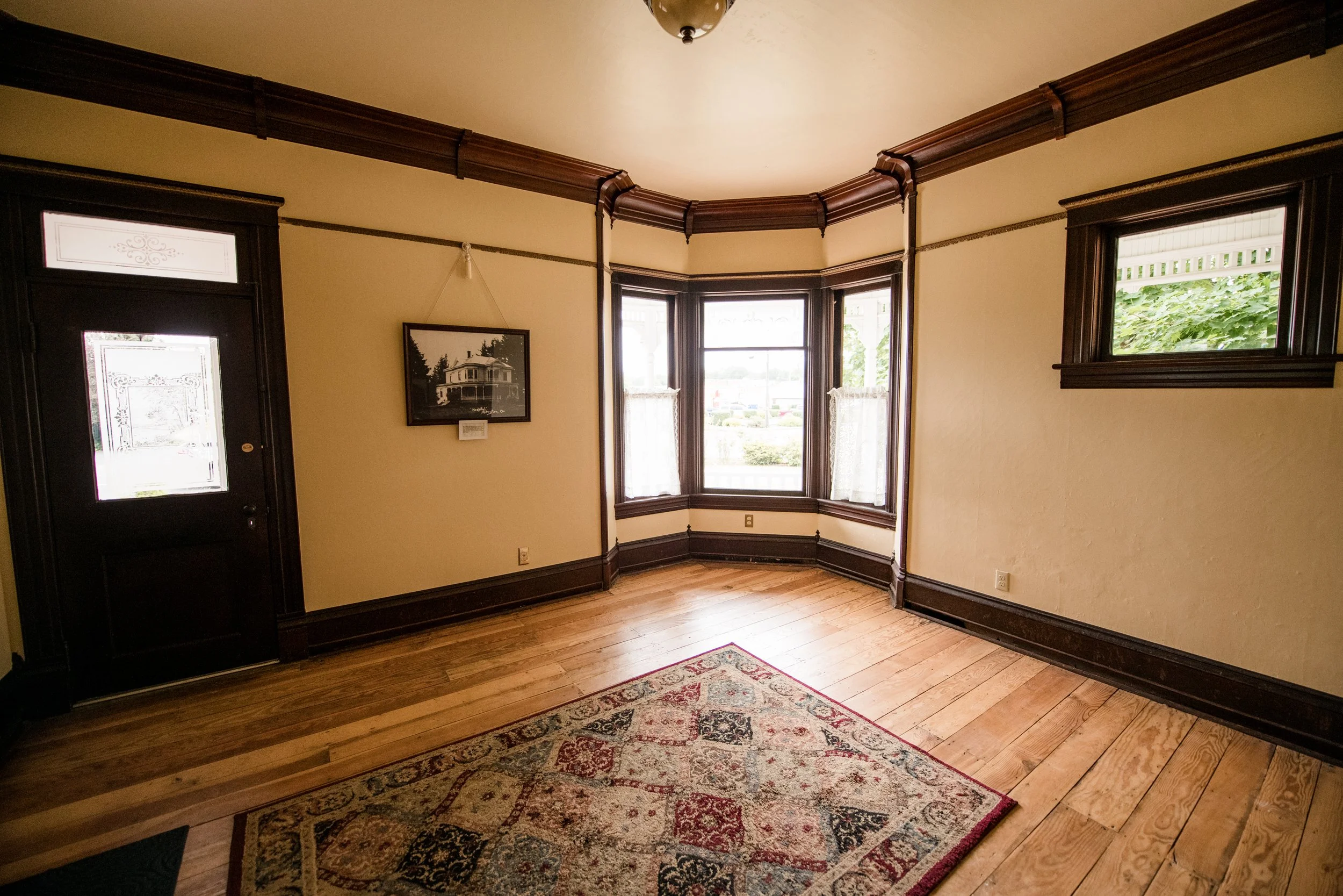The south parlor of the 1903 Brown House with a patterned rug, and a wall with three windows and a framed picture of Stayton's first hospital, decorated with dark wood trim and cream-colored walls.
