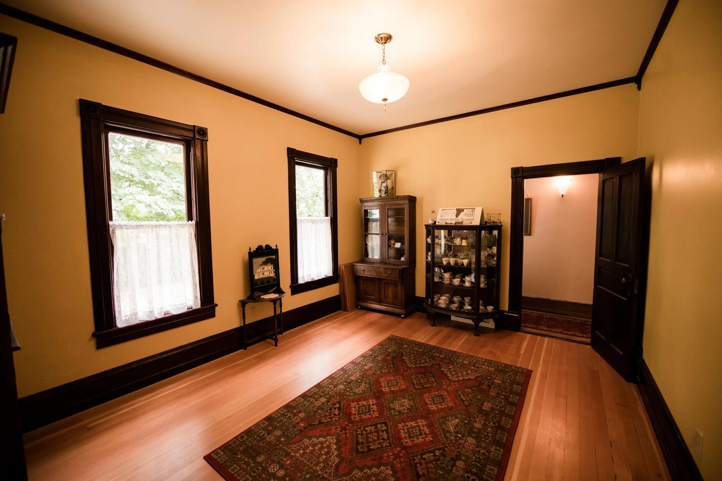 A corner of the dining room of the Brown House with yellow walls, dark wood trim, two windows with lace curtains, a wooden cabinet, and a glass display case with teacups. Ready for any event.