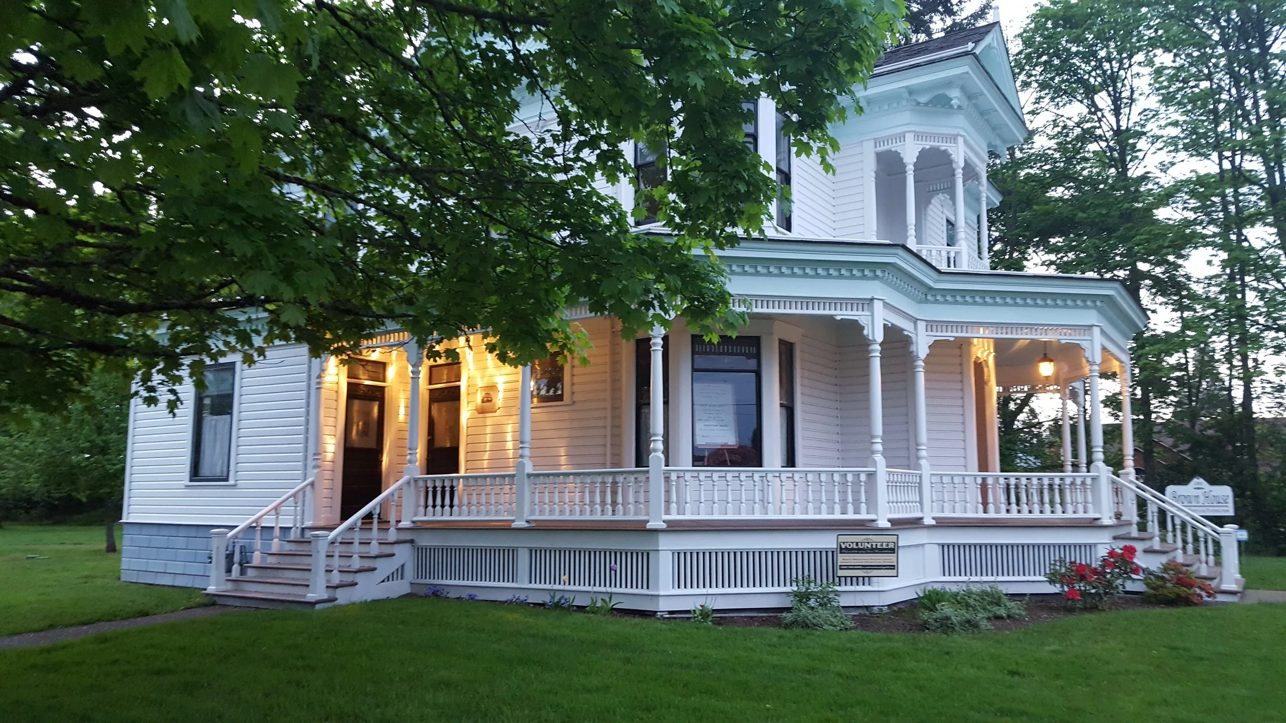 The 1903 Brown House in Stayton, Oregon with a wraparound porch surrounded by trees and a well-maintained lawn, with some exterior lighting on at dusk.