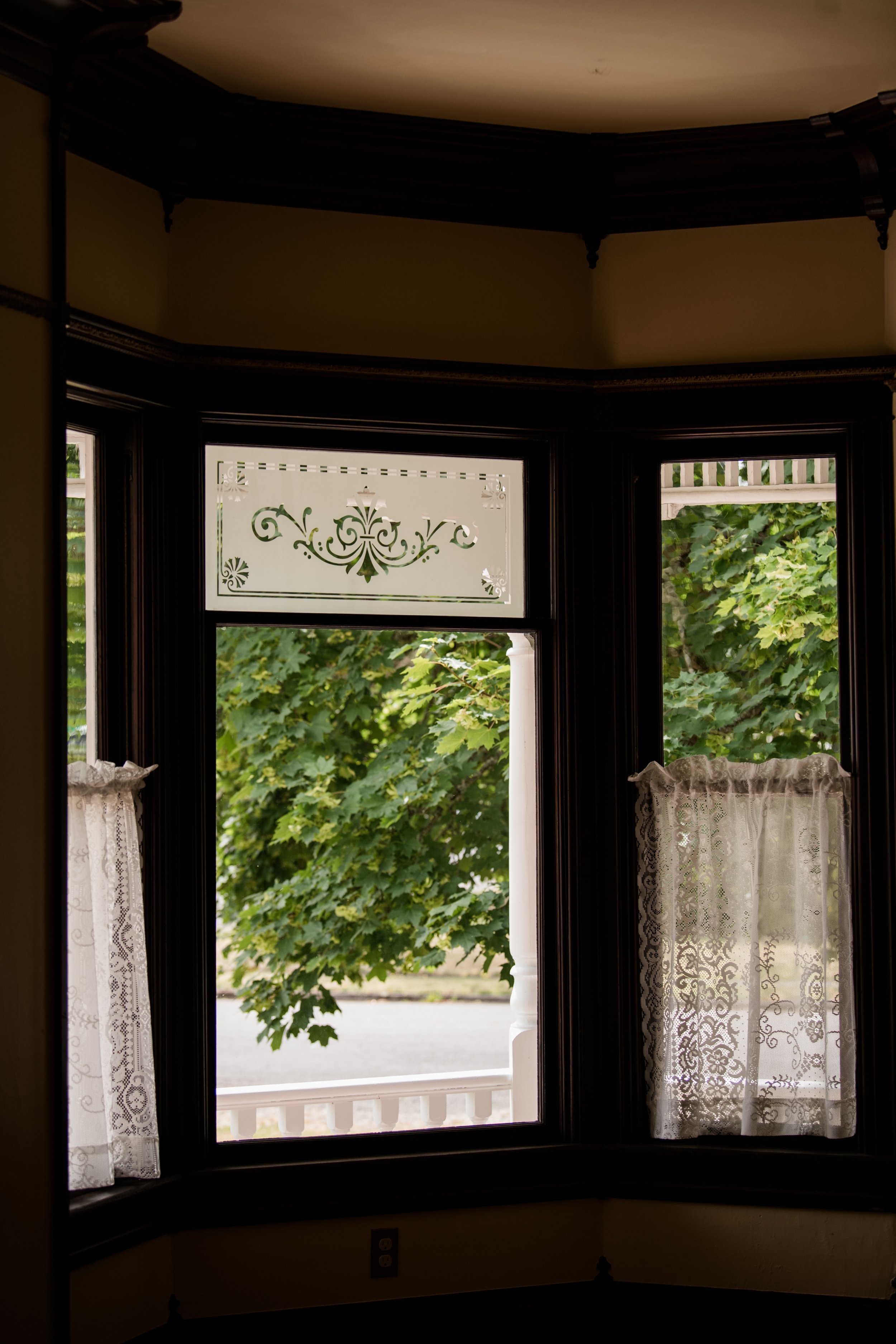 View from a large bay window in the 1903 Brown House in Stayton, Oregon with intricate stained glass detail at the top, framed by lace curtains, overlooking green leafy trees outside.
