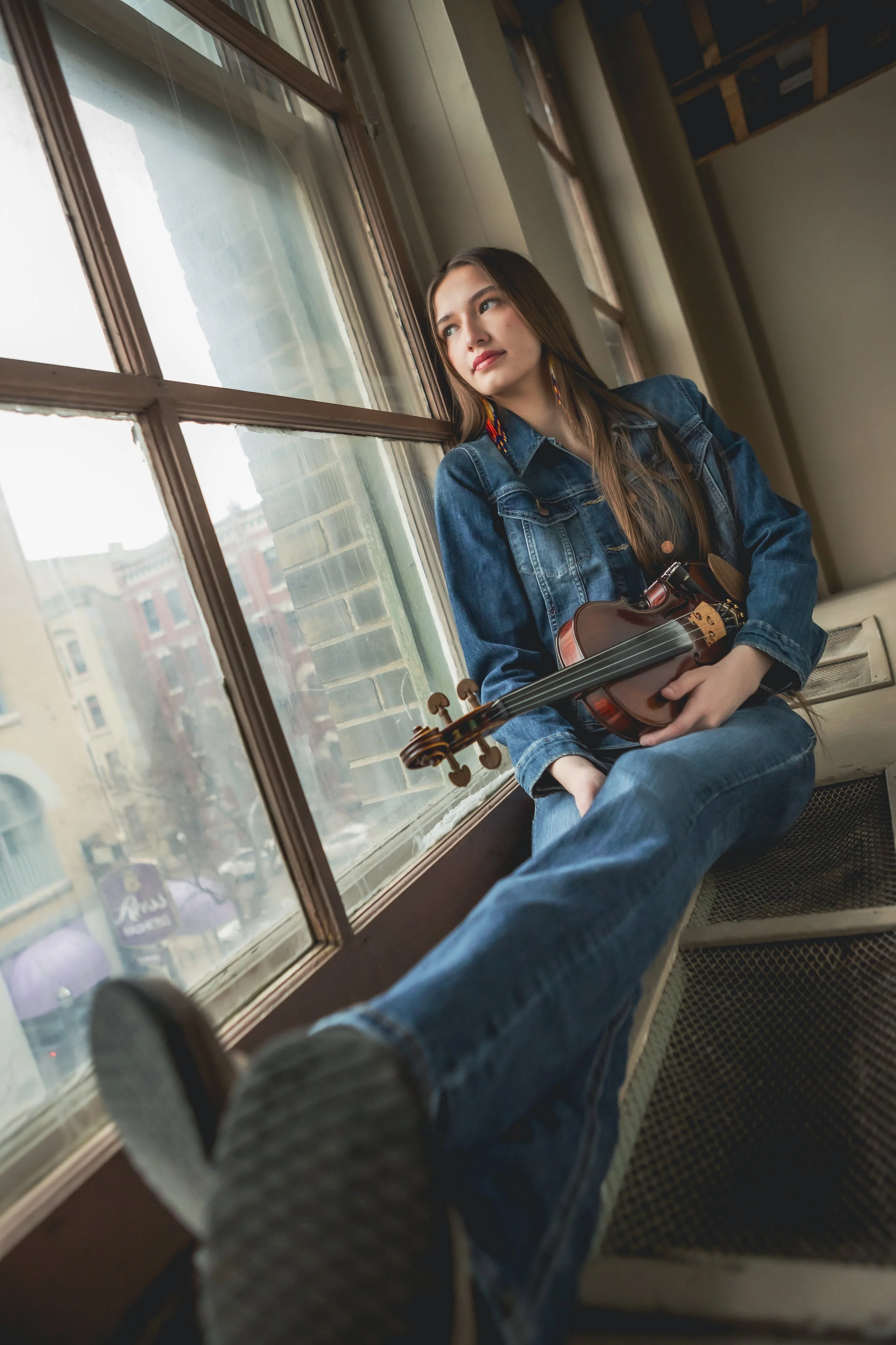 A young woman sitting by a large window, holding a violin, gazing outside, with a cityscape visible through the window.