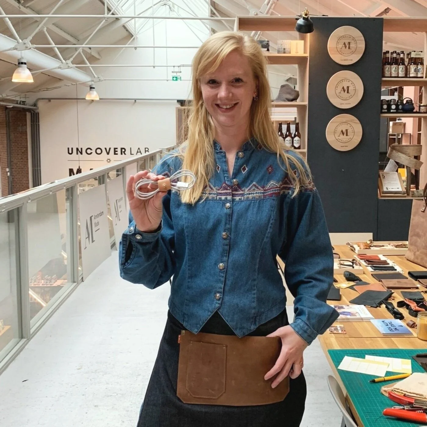 Vrouw met lang blond haar die glimlachend een gekleurd kabel vastheeft en een apron draagt in een ambachtelijke werkruimte met houtbewerking en producten op de achtergrond.