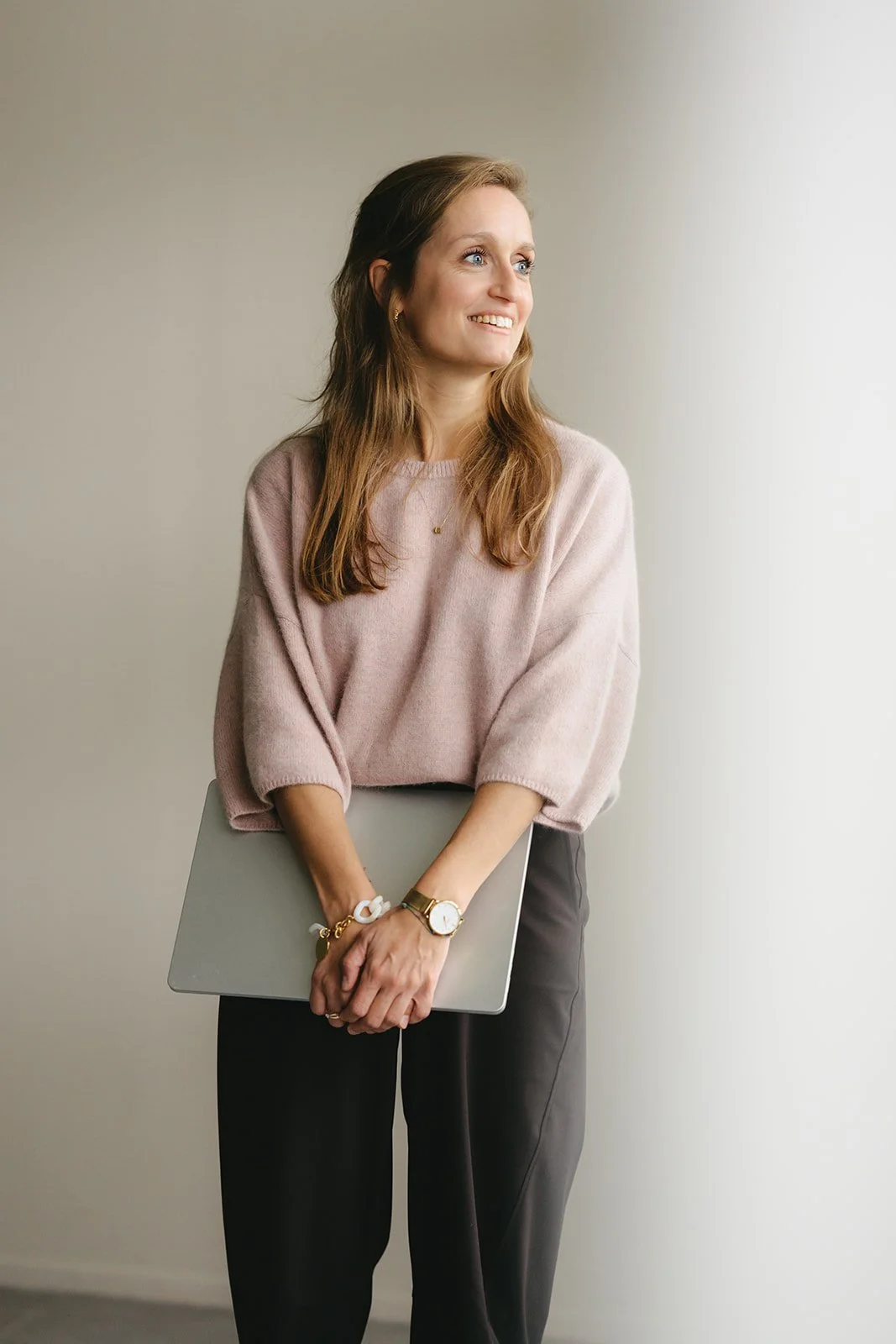 Vrouw met bruin haar, roze trui, zwarte broek, houdt een laptop vast, glimlacht, staand voor een lichtgekleurde muur.