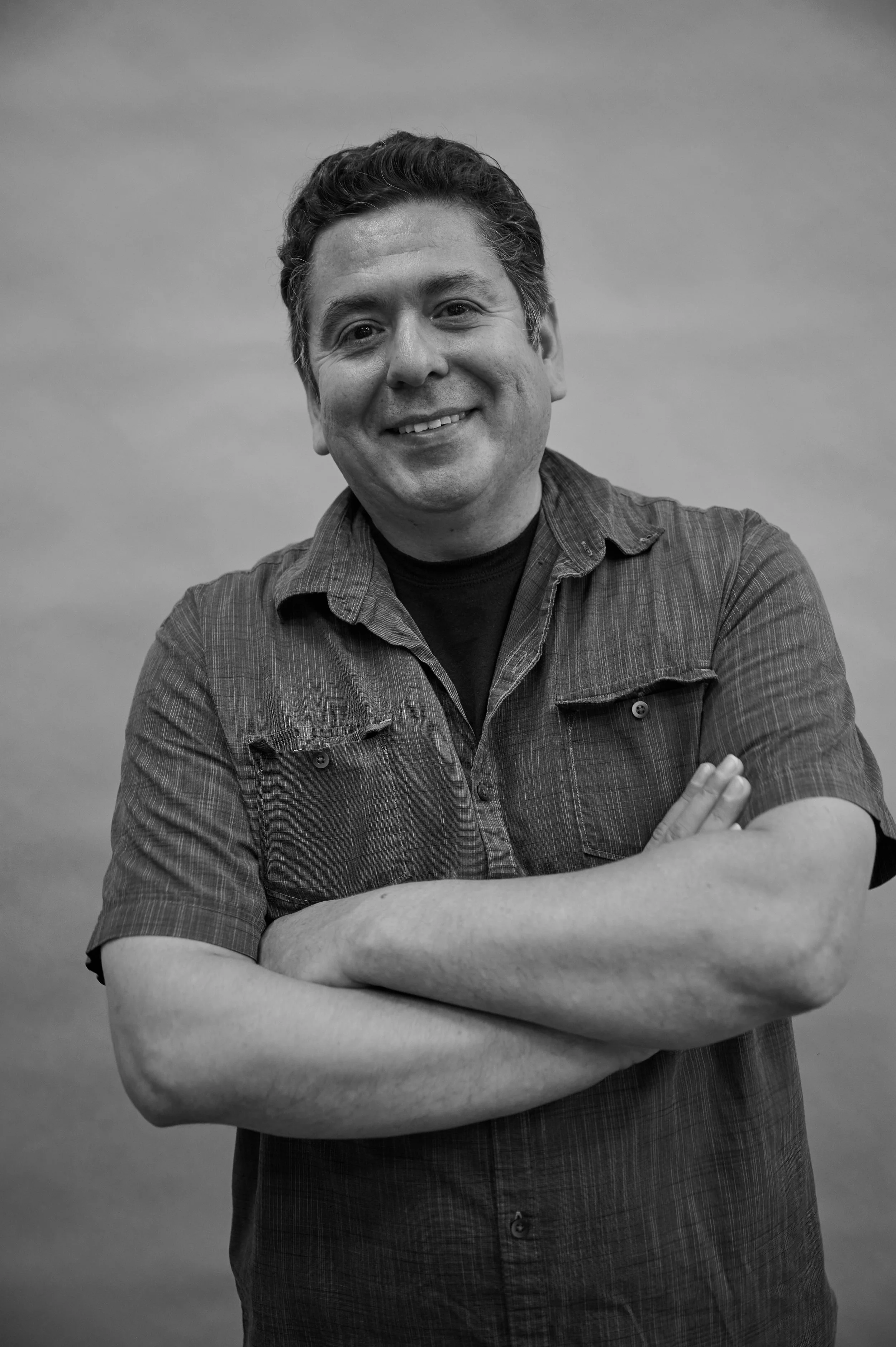 Black and white photograph of a man smiling with arms crossed, wearing a short-sleeved button-up shirt, standing against a plain background.
