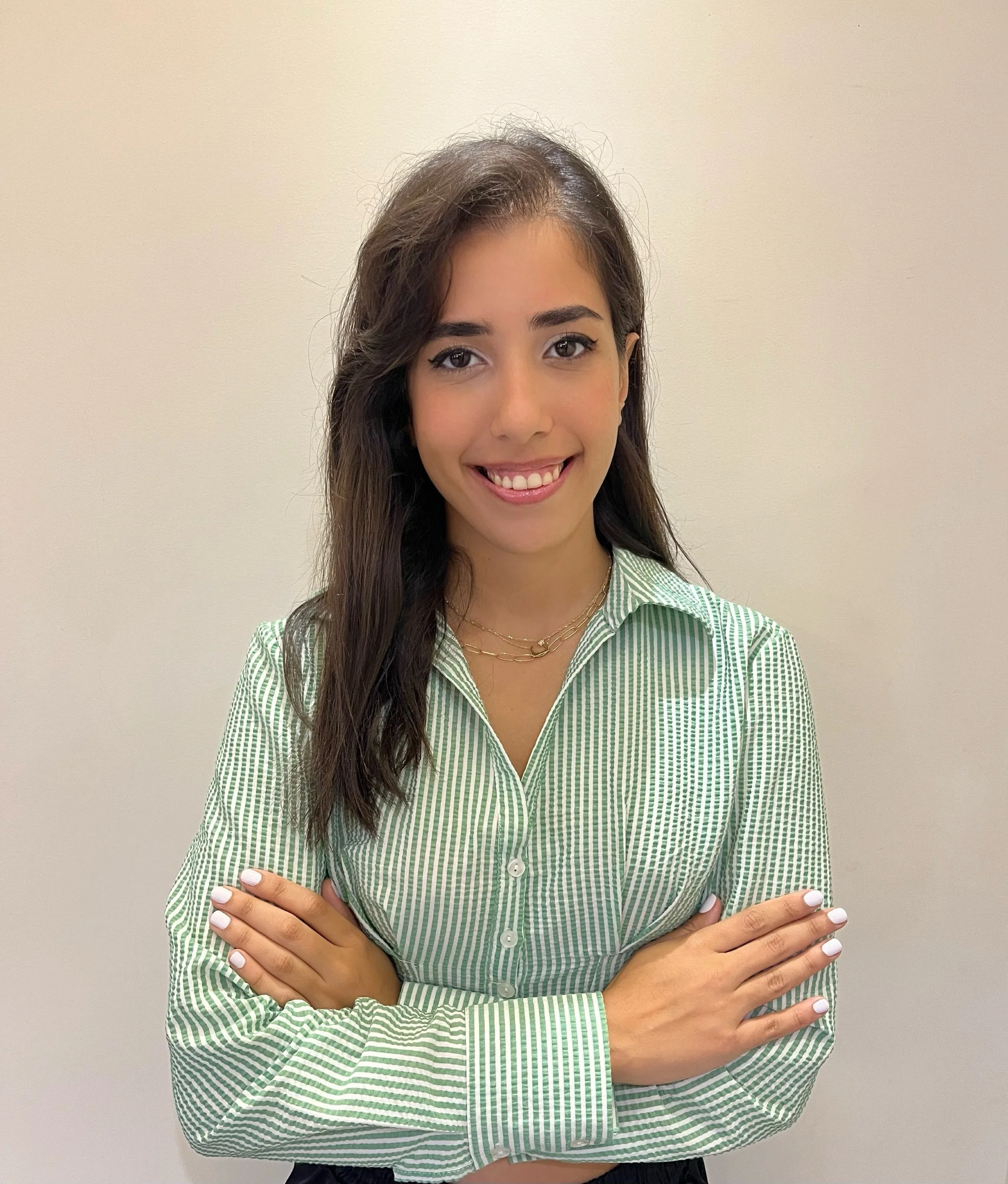 A young woman with long dark hair smiling with crossed arms, wearing a green striped button-up shirt and gold jewelry, standing against a plain beige wall.