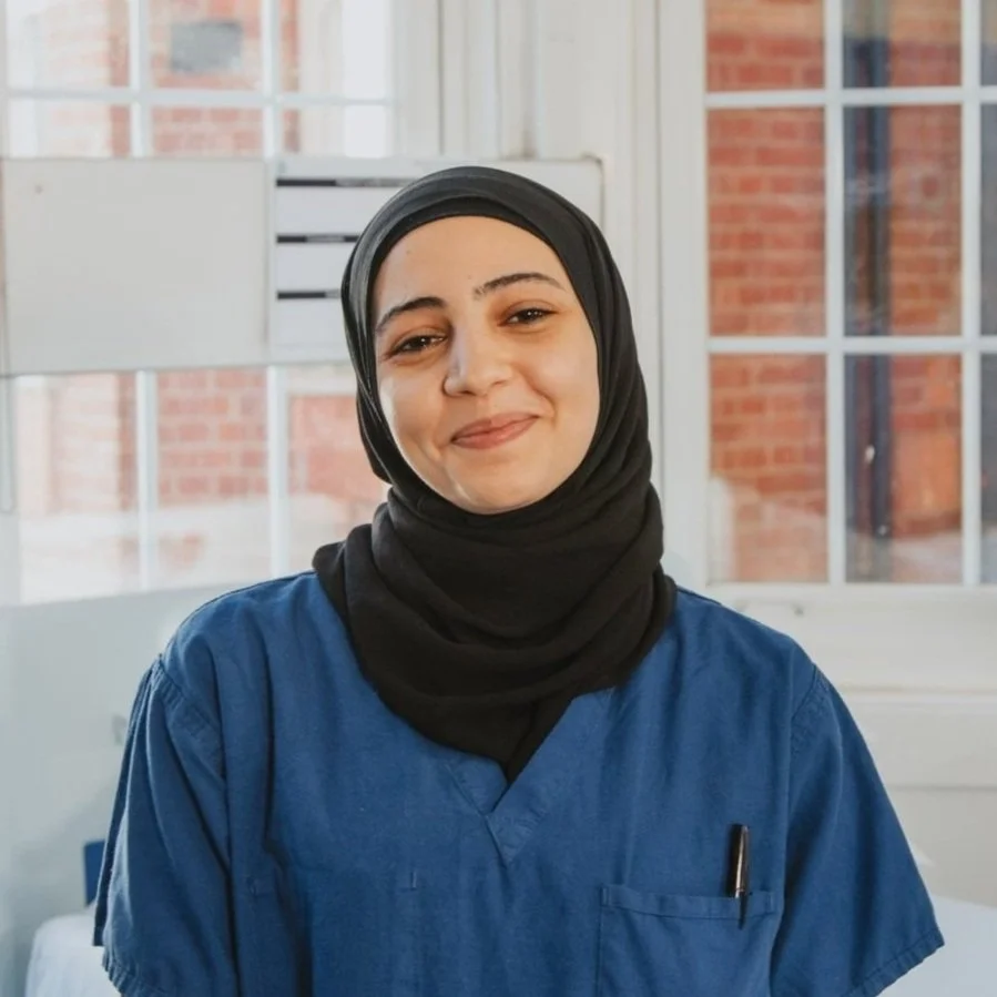 A woman in blue scrubs and a black hijab smiling indoors with a window showing a brick building outside.