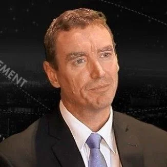 A man in a suit and tie with a serious expression, against a dark background.