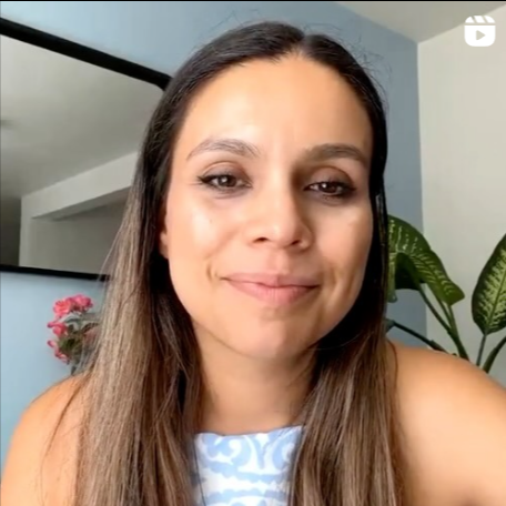 Close-up of a woman with long brown hair and light makeup, smiling in a room with a plant and a mirror behind her.
