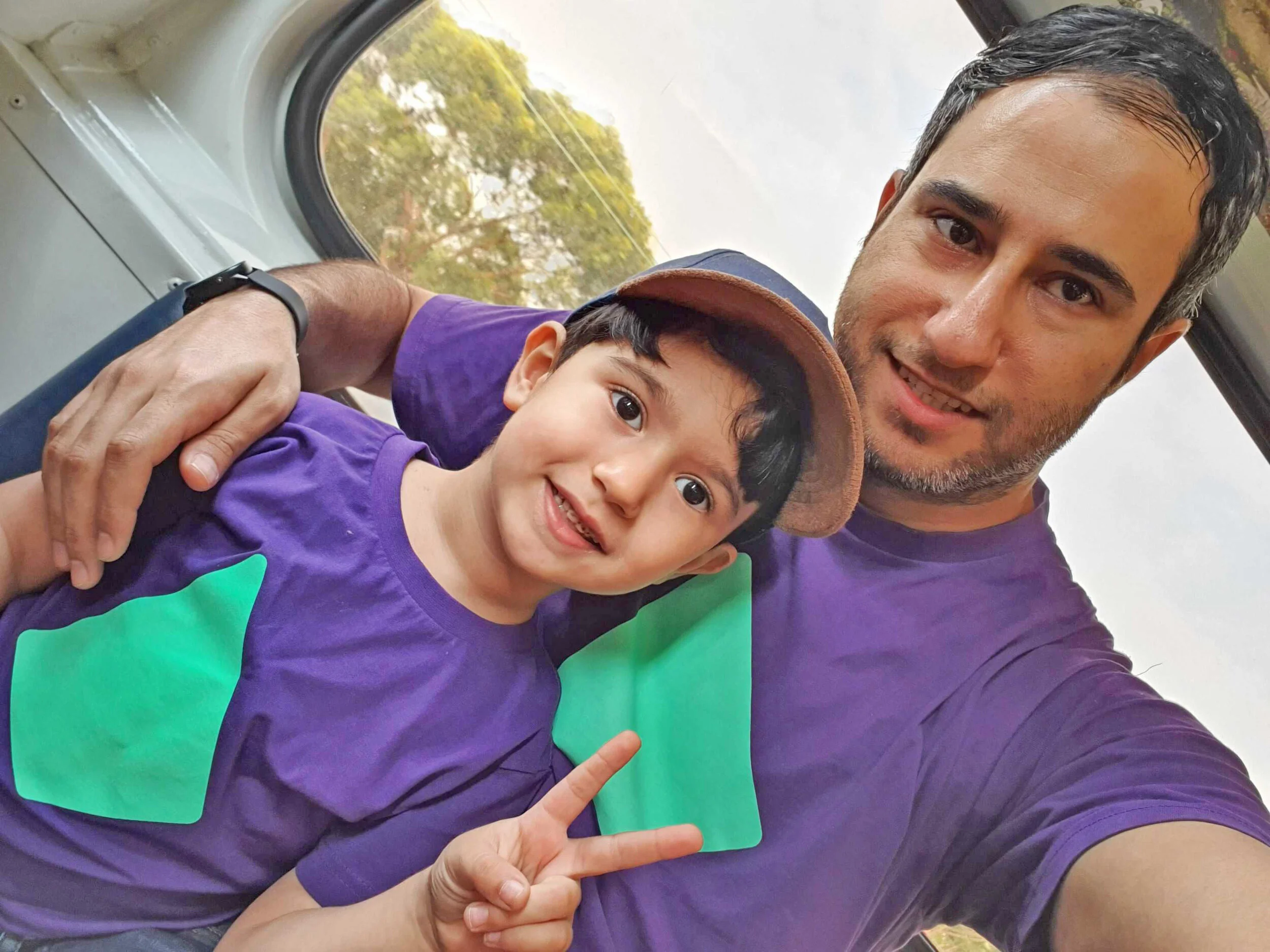 A man and a young boy taking a selfie inside a vehicle. The boy is smiling and making a peace sign, and both are wearing purple shirts with green rectangles on them. The man has short grayish hair and a slight beard, and the boy is wearing a brown hat. The background shows a window with trees outside.