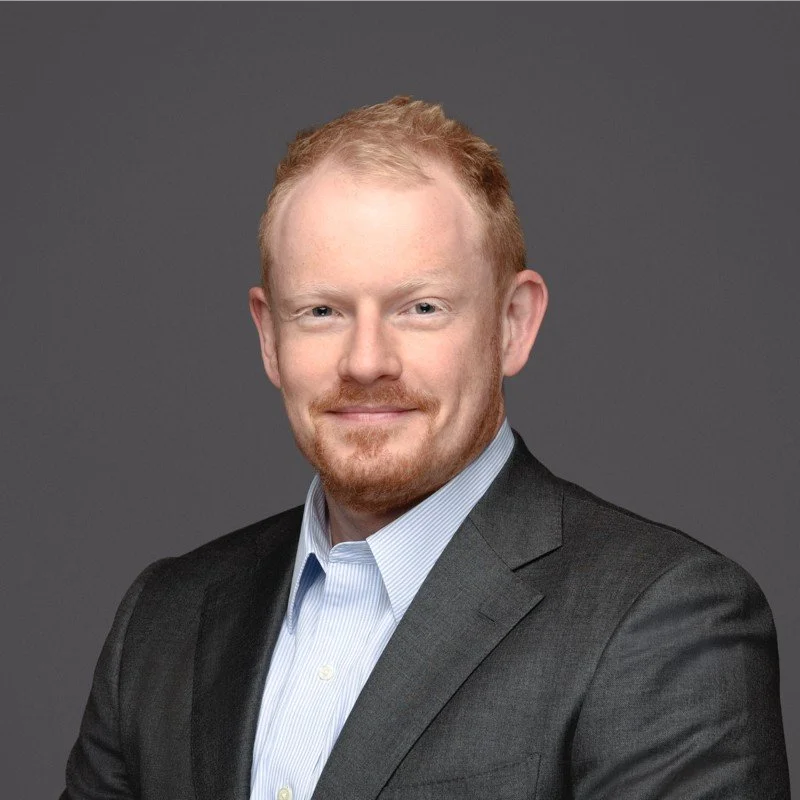 A professional headshot of a man with light skin, red hair, and a beard, wearing a dark suit and light blue shirt, smiling against a gray background.
