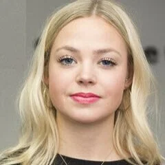 Close-up of a young woman with long blonde hair, light makeup, wearing a black top, looking at the camera.