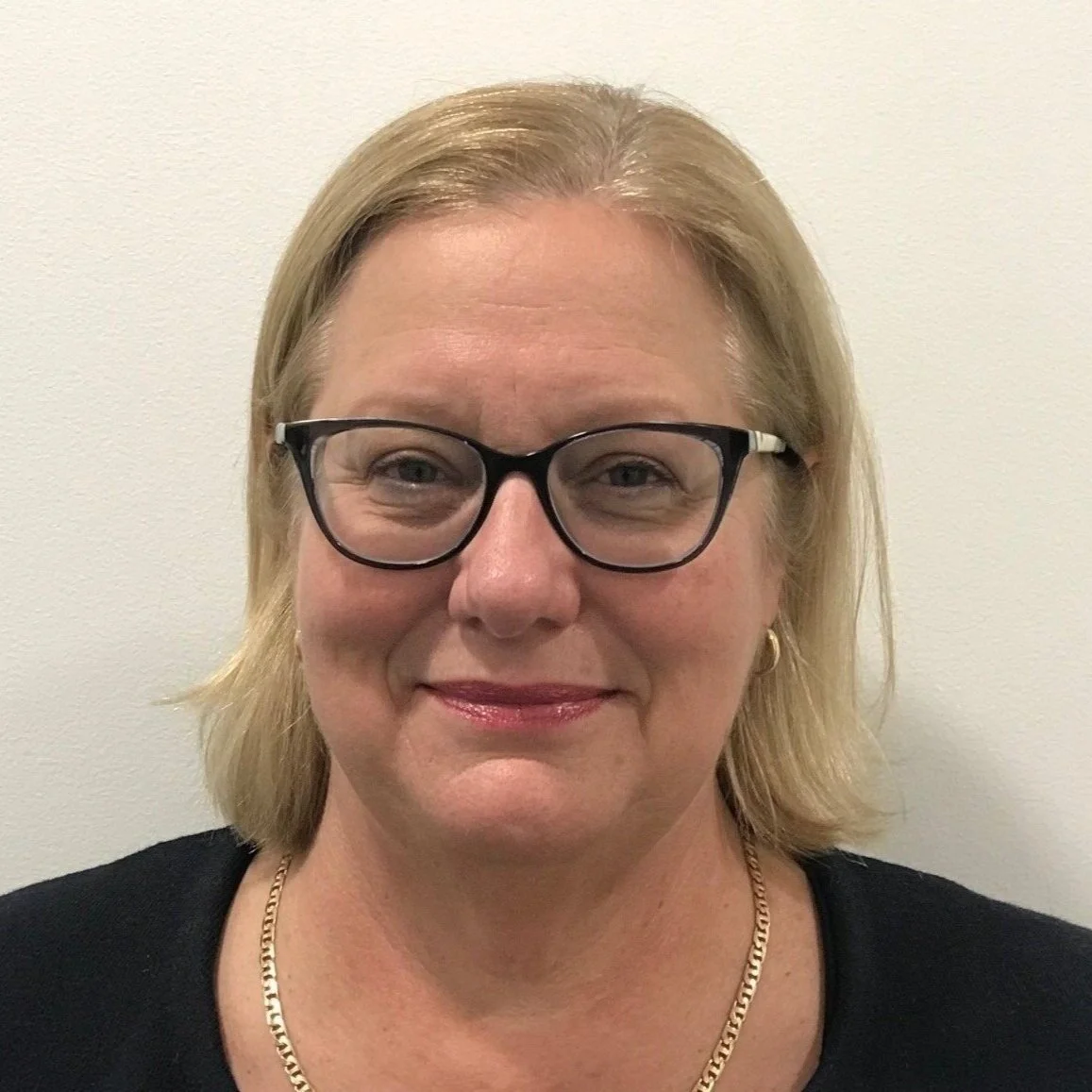 A woman with blonde hair, glasses, and gold jewelry smiling at the camera against a plain white wall.
