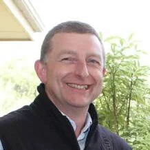 A smiling man with short hair, wearing a black jacket, standing outdoors near a house with a window, green trees, and bright sunlight.