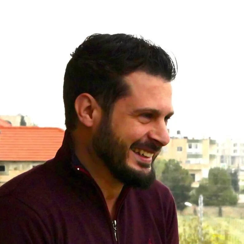 A man with short dark hair and a beard smiling outdoors with buildings and trees in the background.