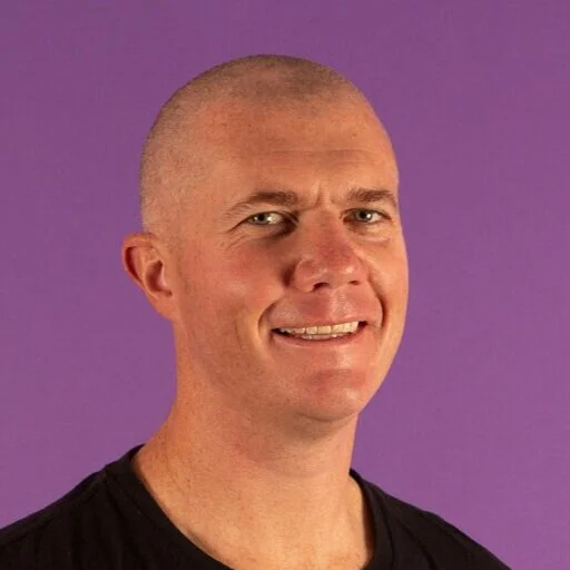 A portrait of a smiling man with a shaved head, wearing a black shirt, against a purple background.