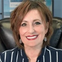 A woman with short, curly, auburn hair smiling, wearing a navy and white striped blouse, sitting in an office chair.