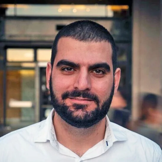 A man with dark hair, a full beard, and a mustache smiling at the camera outside a building
