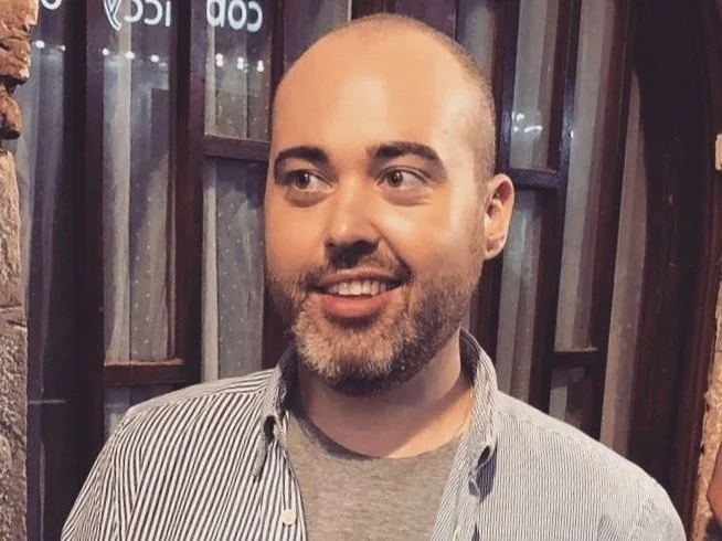 A man with a beard and a shaved head, wearing a gray t-shirt and a striped shirt, smiling and looking to the side in a setting with wooden and metal surroundings.