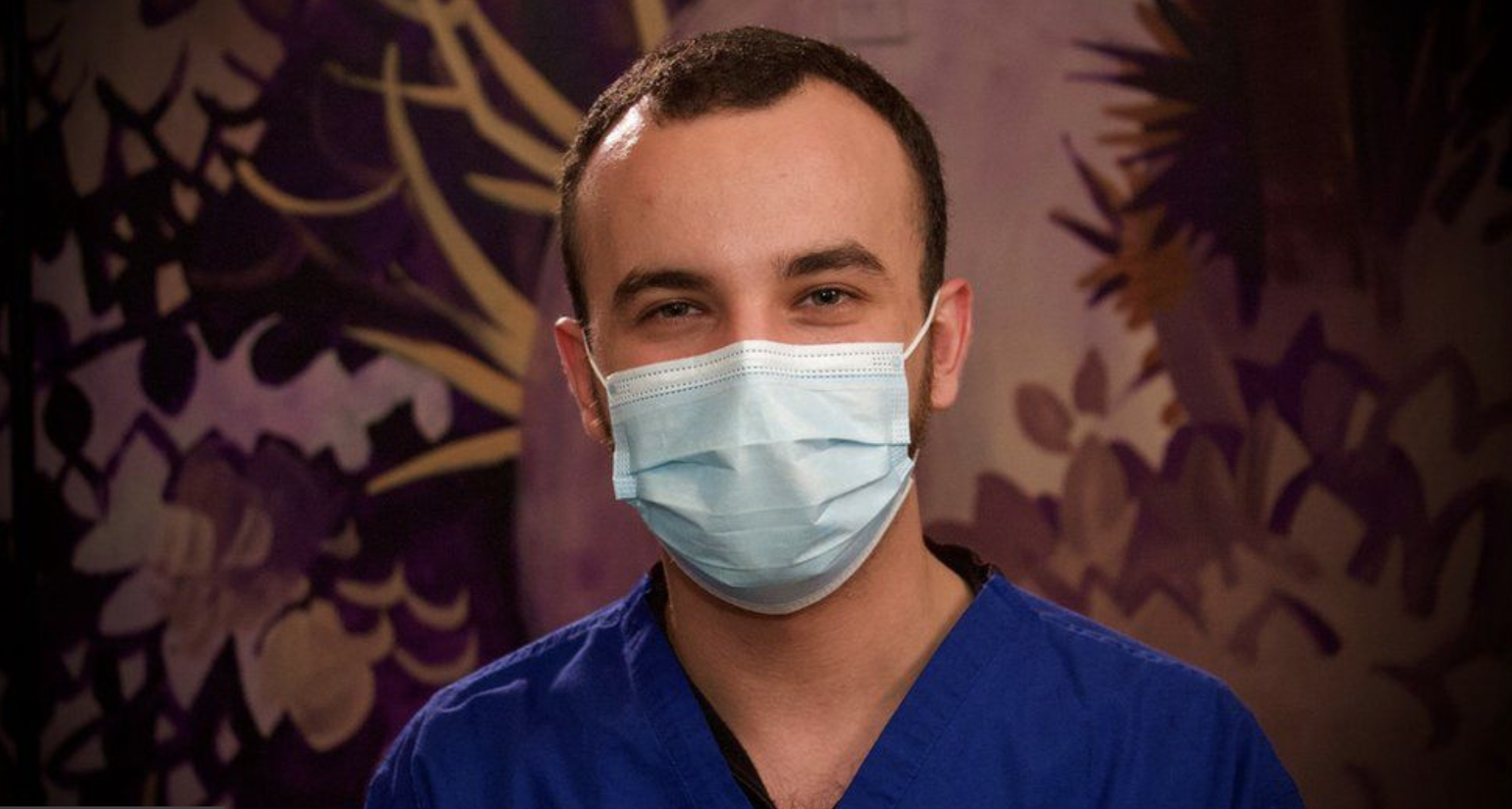 A man wearing blue medical scrubs and a light blue face mask, smiling at the camera, in front of a dark purple background with gold and purple leaf patterns.