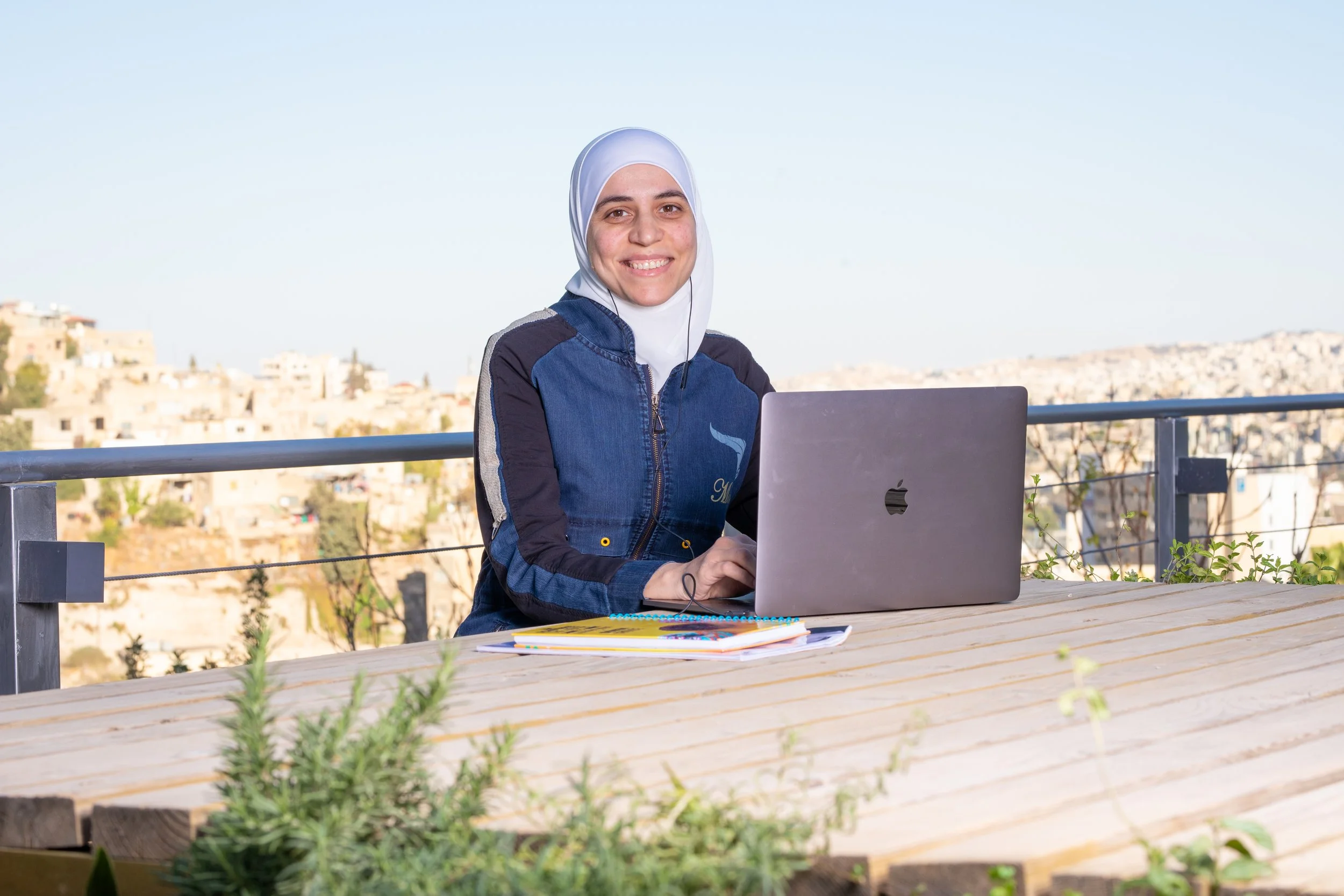 Young woman wearing a white hijab and a denim jacket sitting outside at a wooden table with a laptop, notebooks, and pens, smiling at the camera, with a cityscape in the background.