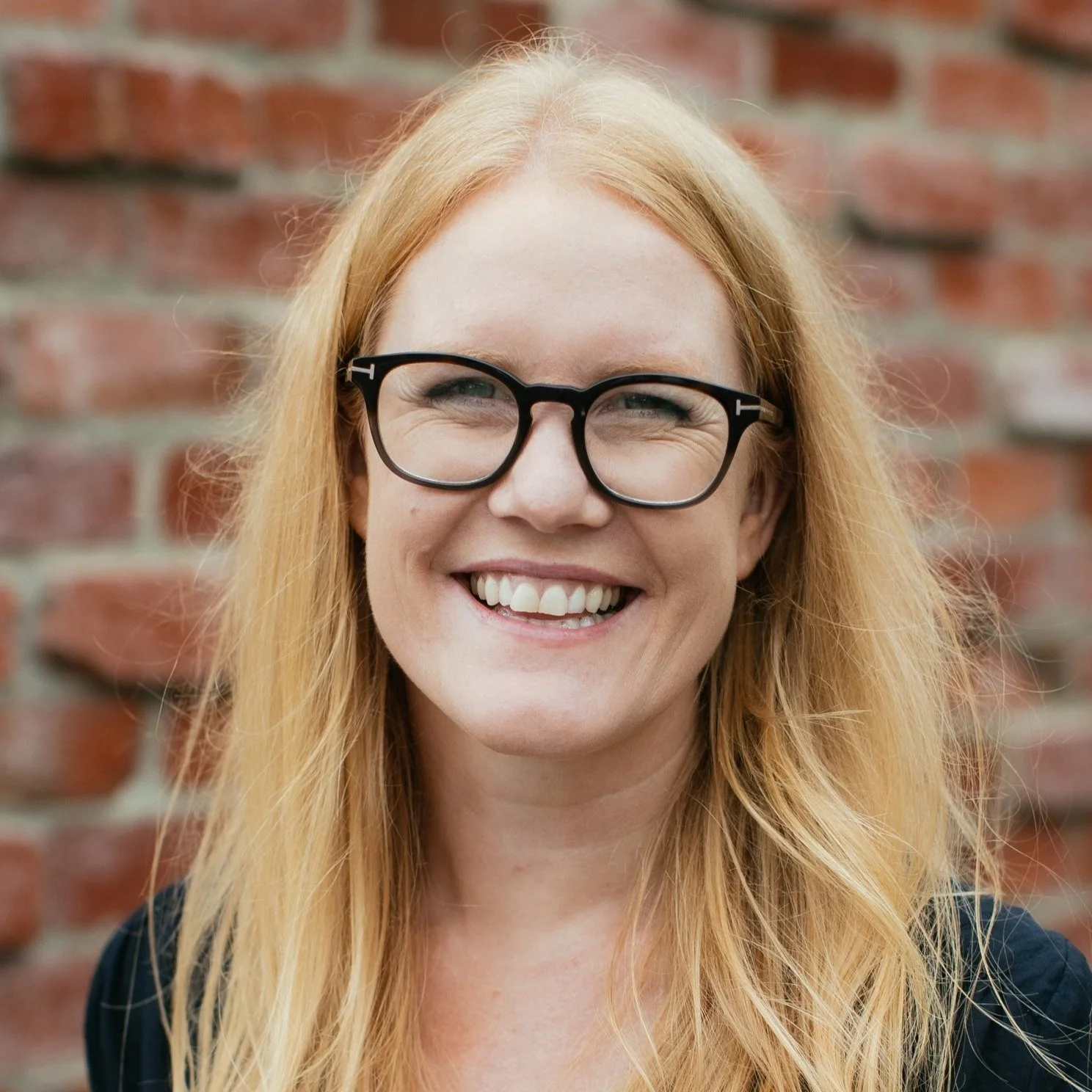 Close-up of a smiling woman with long red hair, wearing black-rimmed glasses, standing in front of a brick wall.