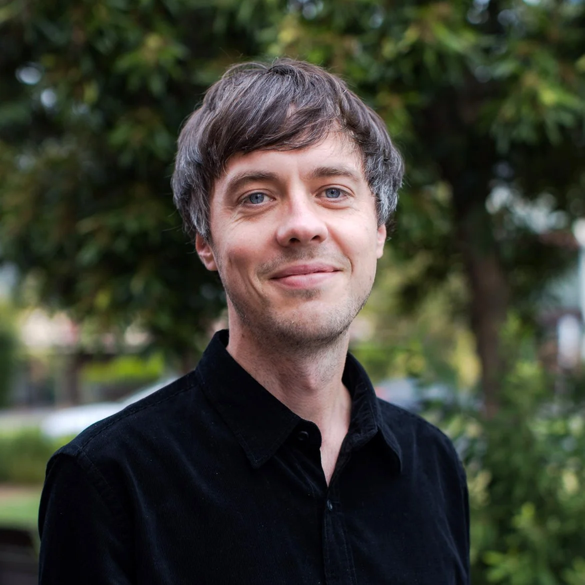 A man with grayish-brown hair and blue eyes, smiling, wearing a black shirt outdoors with trees in the background.