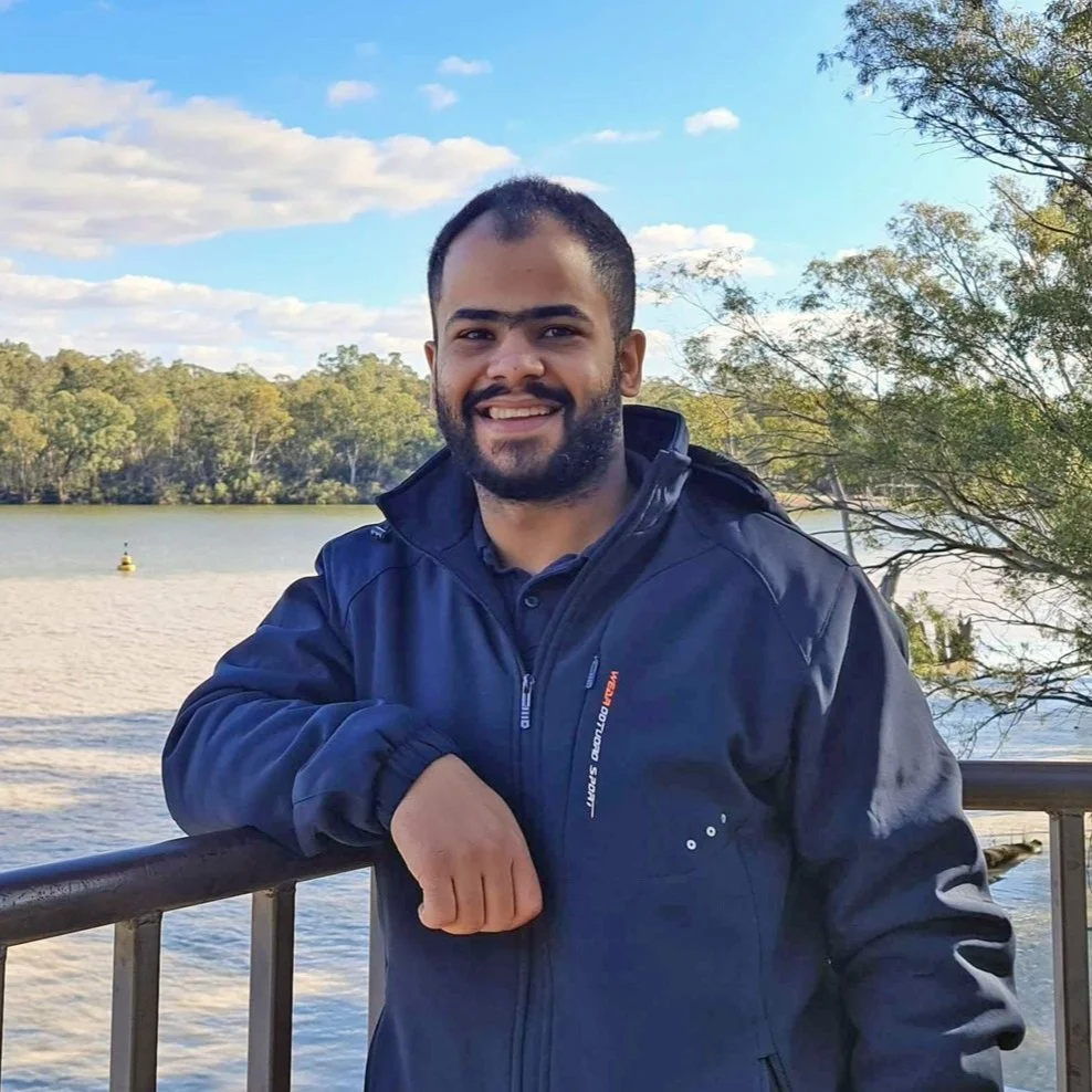 A man with a beard and short dark hair smiling, standing outdoors near a body of water with trees in the background, on a sunny day.