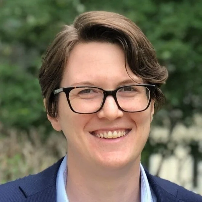 A smiling person with short brown hair, wearing black-rimmed glasses, a blue blazer, and a light blue shirt, standing outdoors with blurred green foliage in the background.