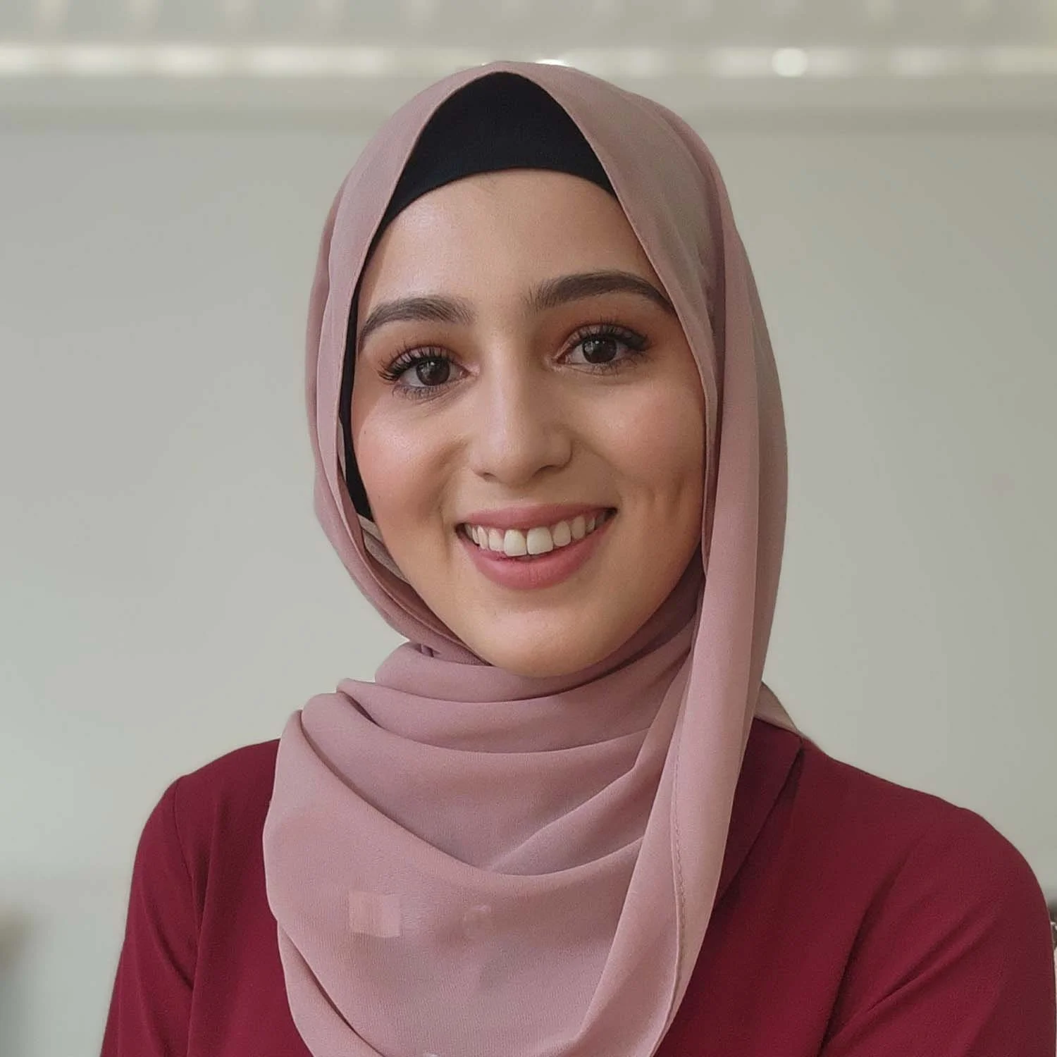 A young woman wearing a pink hijab and maroon top, smiling at the camera against a plain background.