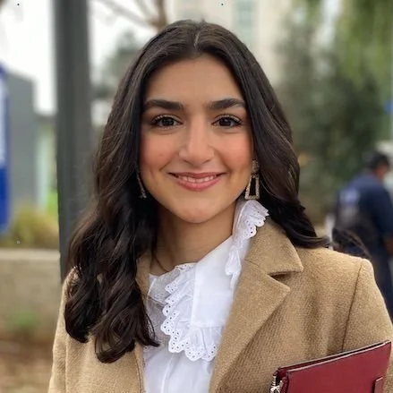 Young woman with dark hair smiling outdoors, wearing a tan blazer and white blouse with lace collar, holding a red folder.