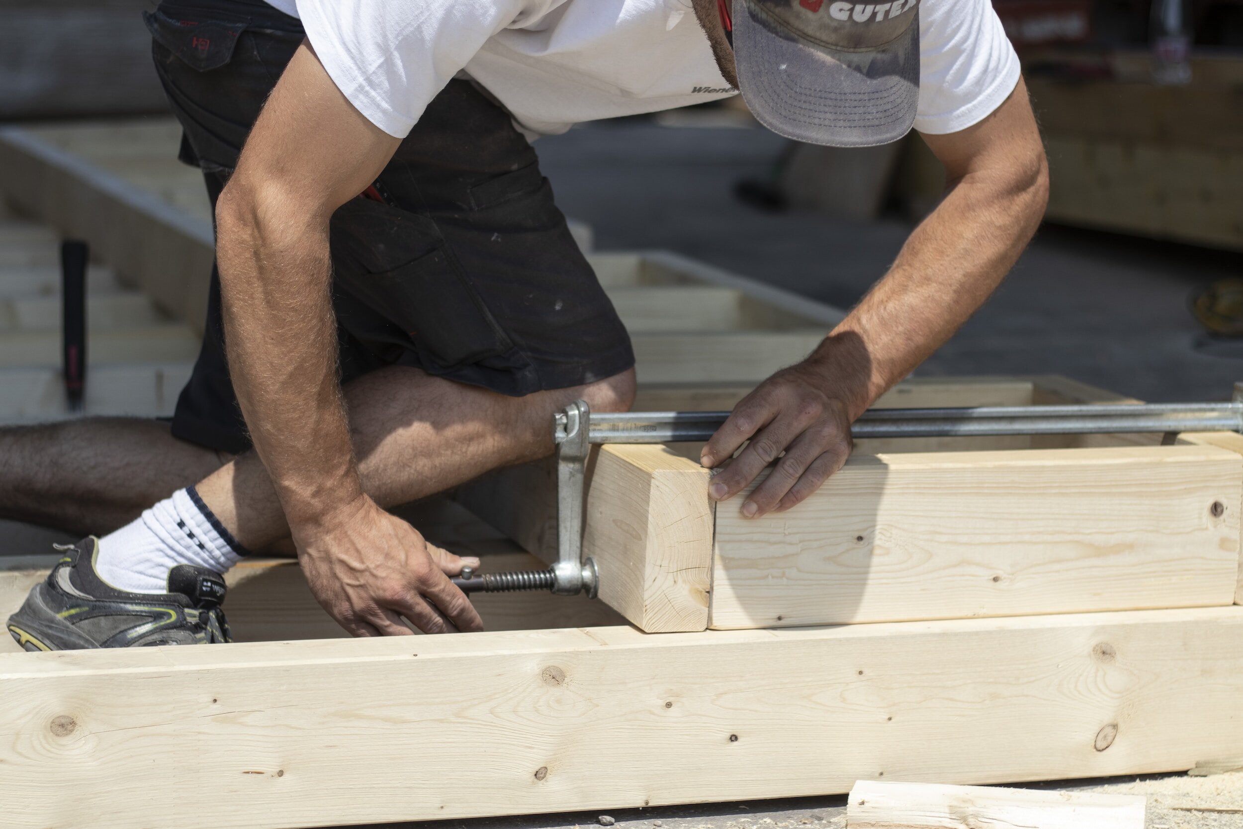 A man is working on a woodworking project, using a clamp to hold a piece of wood in place while assembling a frame or structure outdoors.