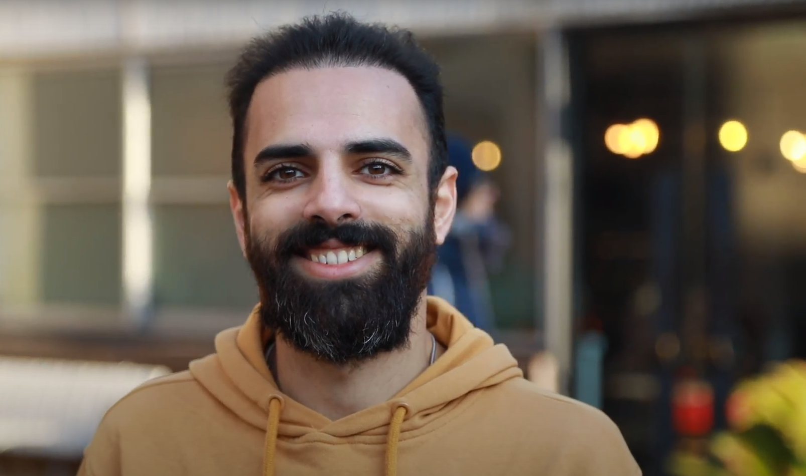 A smiling man with dark hair, a beard, wearing a tan hoodie, standing outdoors in front of a building.