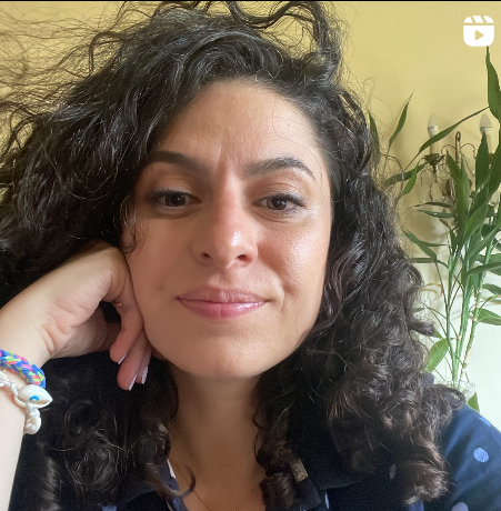 Close-up of a woman with curly dark hair, resting her chin on her hand, wearing a blue shirt and colorful bracelets, with a green plant in the background.