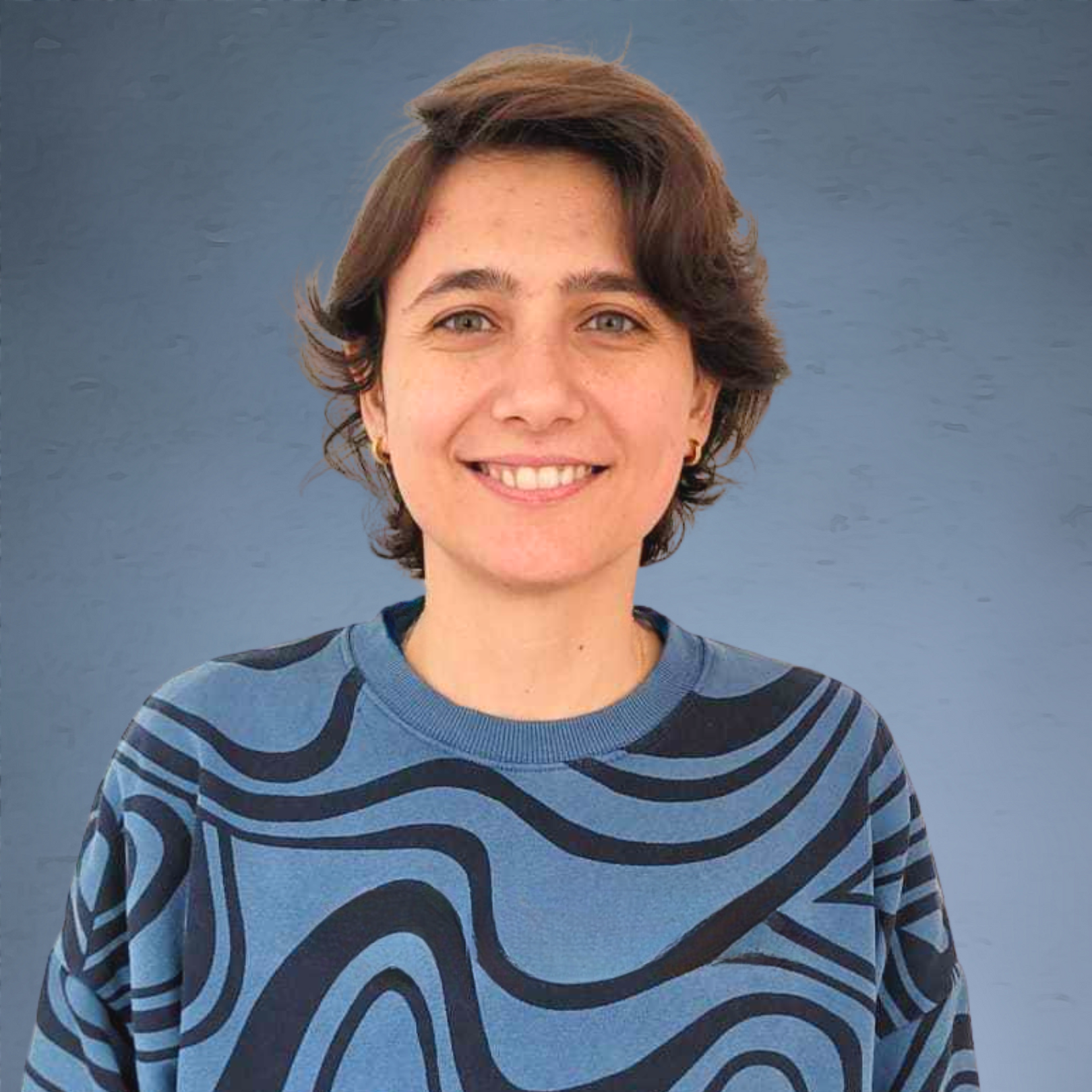 A woman with short brown hair smiling in front of a blue background, wearing a blue and black patterned sweater.